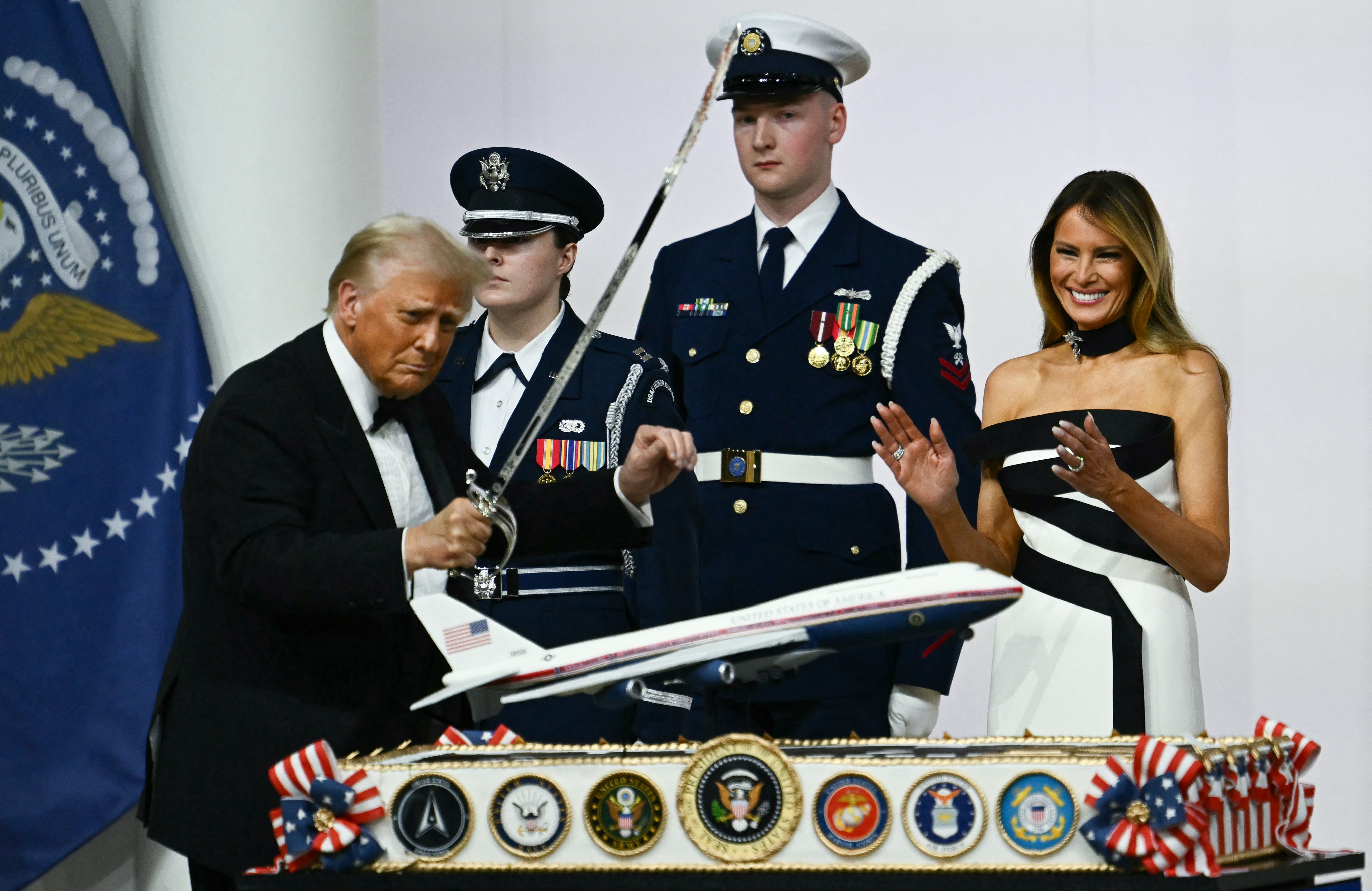 Donald Trump raising a saber to cut a cake with a model of the new Air Force One plane on it. Melania Trump laughs as she watches on. Two members of the military stand behind them.
