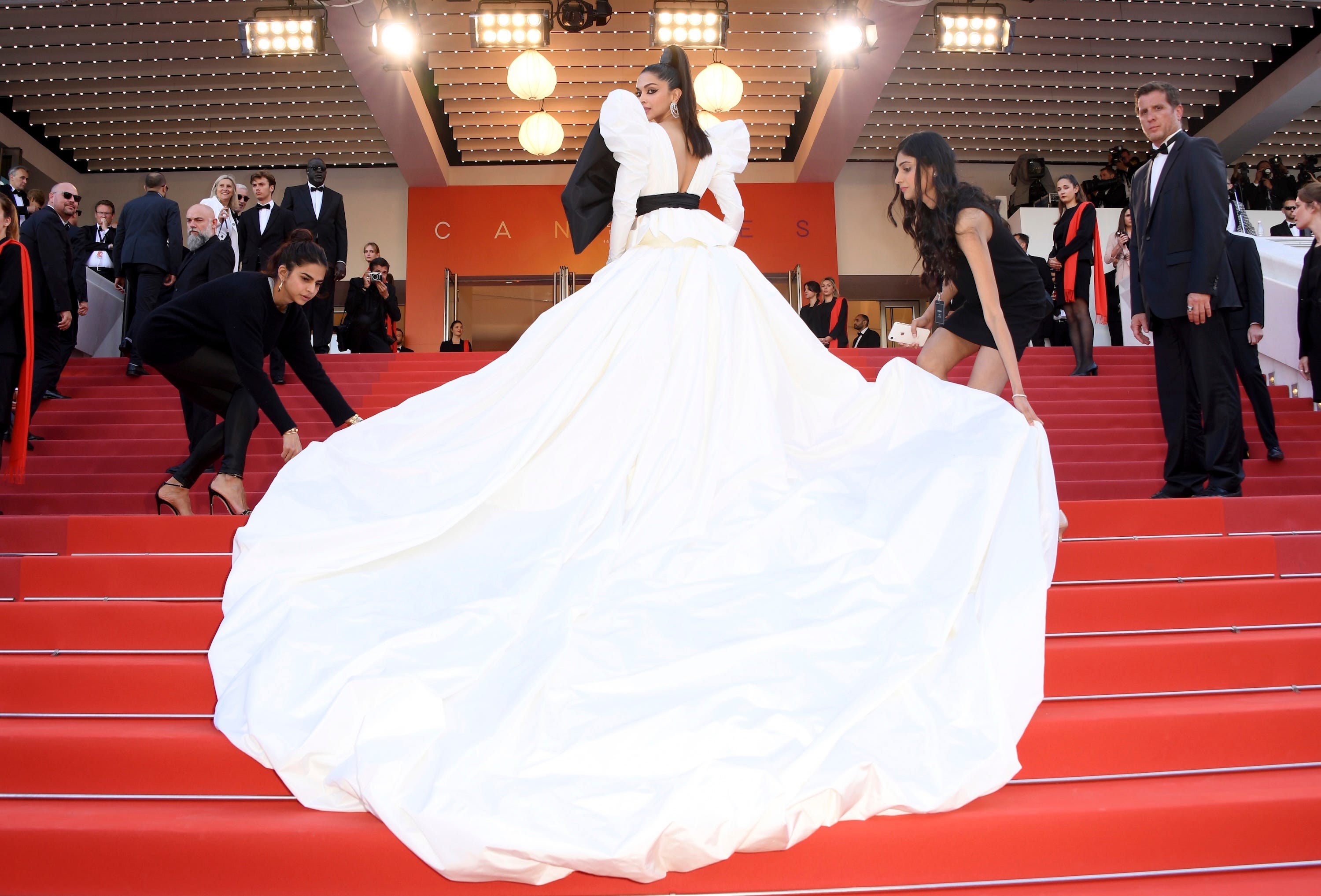 Deepika Padukone poses at the top of steps at the Cannes Film Festival while women adjust the train of her skirt.