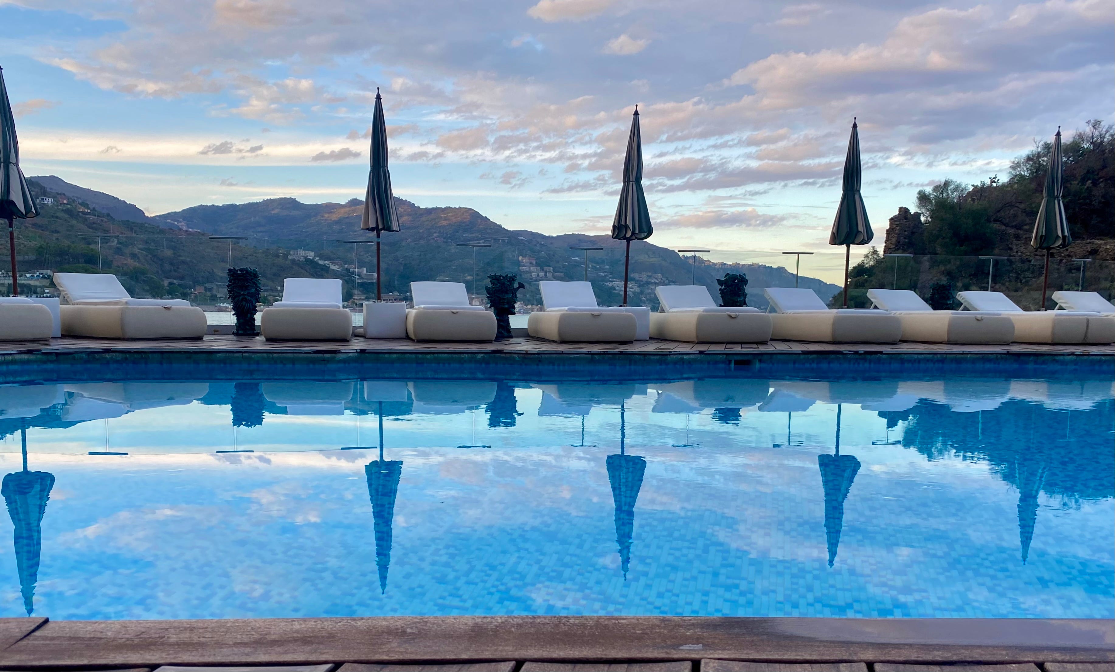 Pool with empty chairs, closed umbrellas next to it reflecting in the pool's blue waters