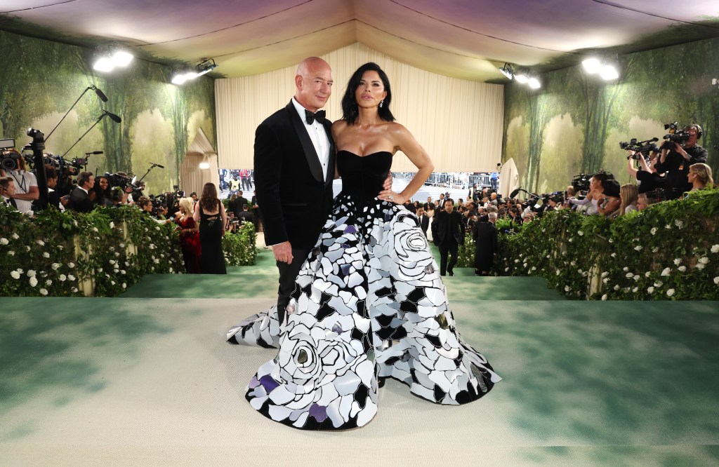 Jeff Bezos in a black tux and Lauren Sanchez in a strapless gown with black top and large black and white skirt with patterns of what appear to be roses on it attend The 2024 Met Gala press can be seen flanking the sides of the background of the image