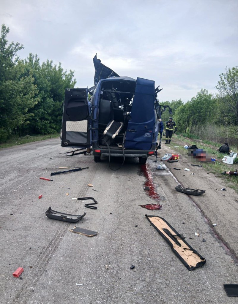 Bus seen after drone strike in Ukraine, bodies blurred, blood seen along road, and debris scattered around road.