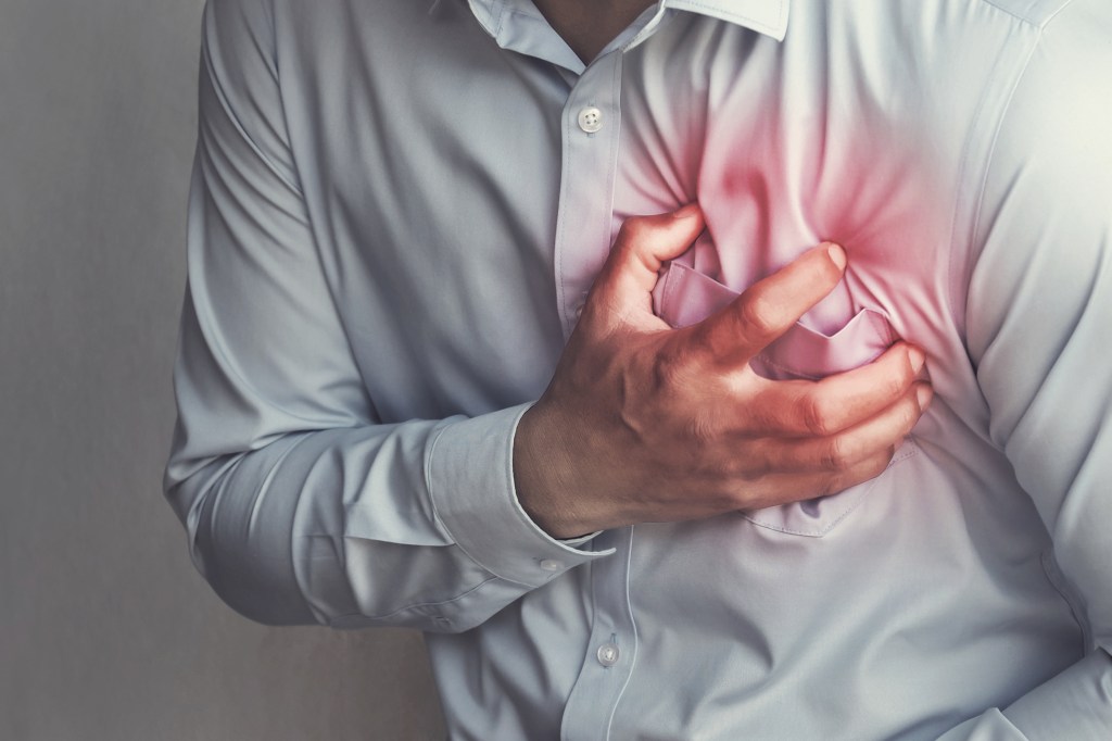 Man in button down shirt clutching chest in pain from a heart attack, illustrated with red highlighting around heart area.