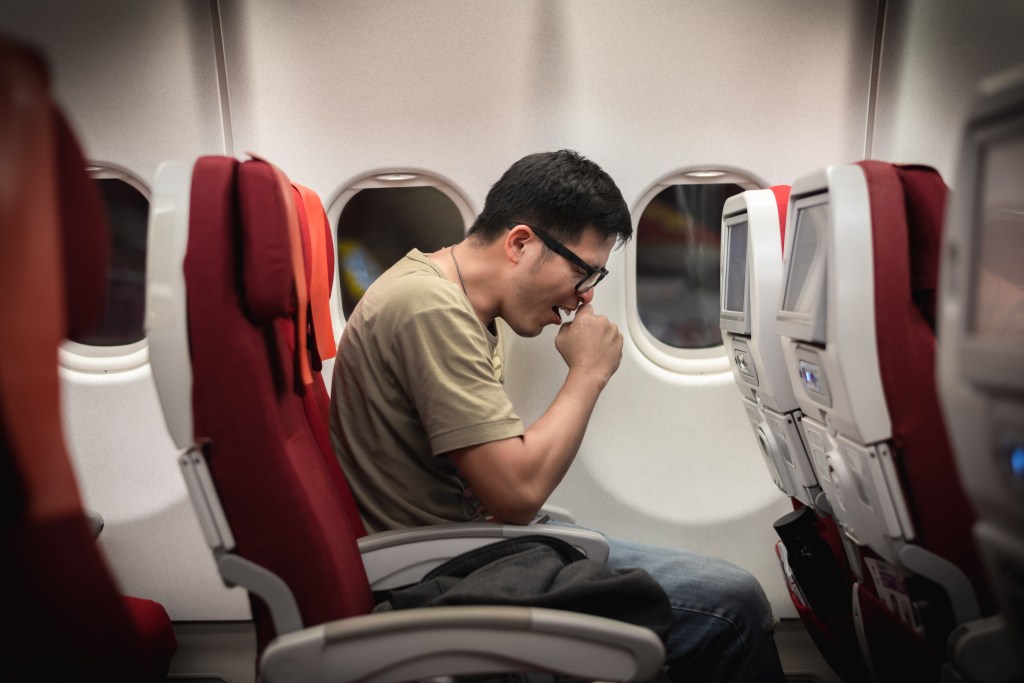 Man appearing unwell while travelling on an airplane with red seats, coughing into hand