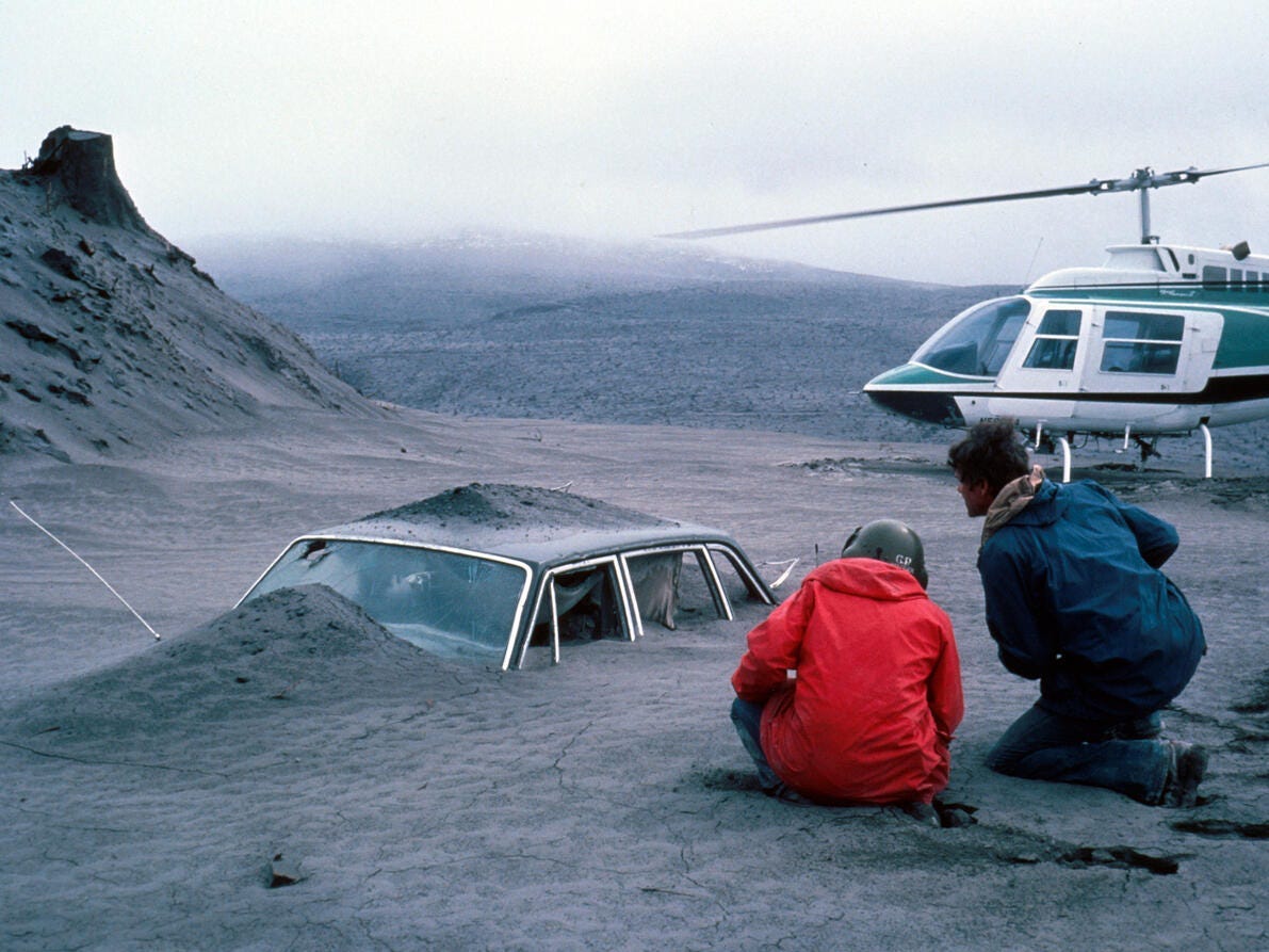 Two people kneel by a car covered in ash by a helicopter