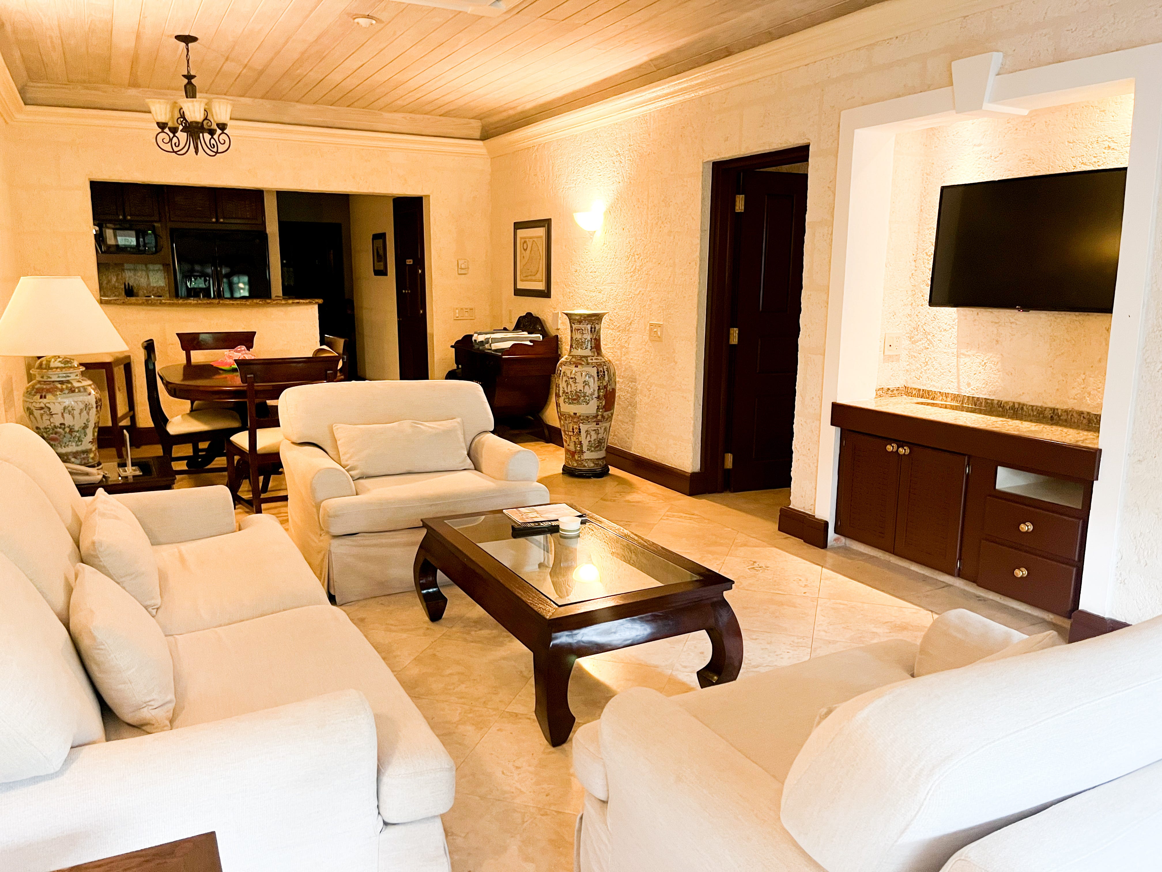 A seating area with white couches, mahogany furniture, and a TV that leads into a dining area.