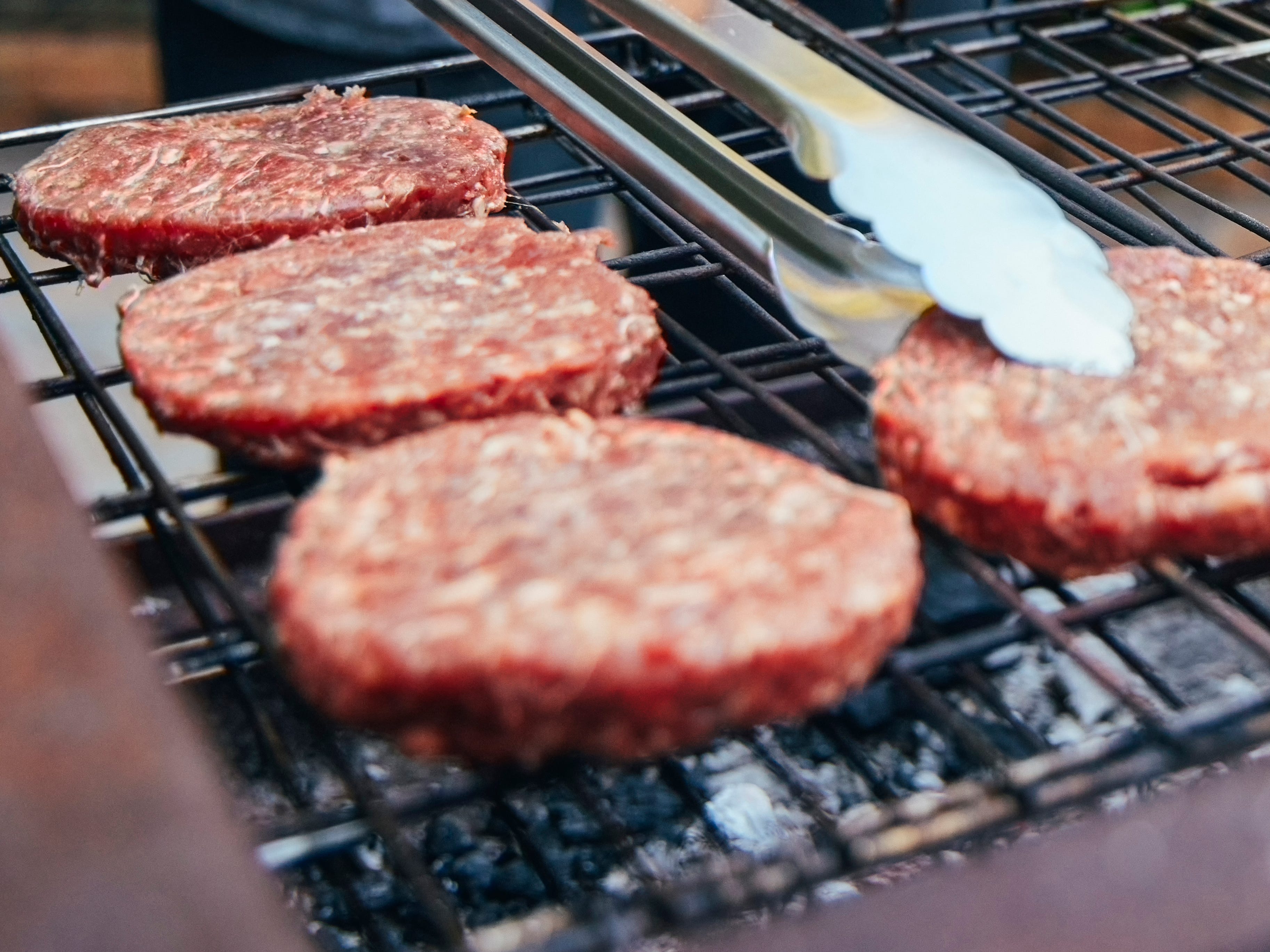 Raw burgers on a fiery grill with tongs about to flip one over