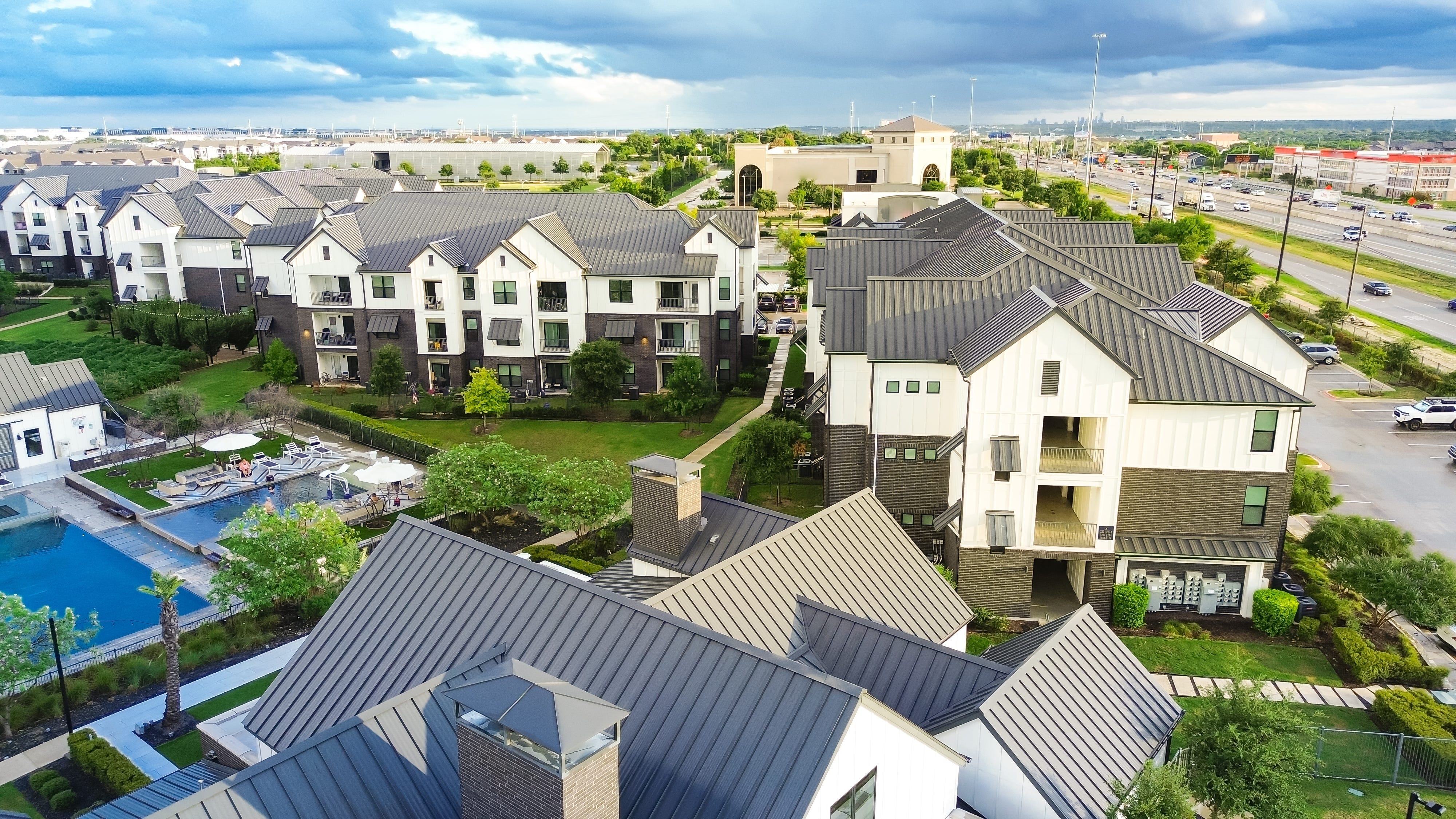 A housing complex with multiple white, gray-roofed buildings surrounding green lawns and a pool sits next to a highway