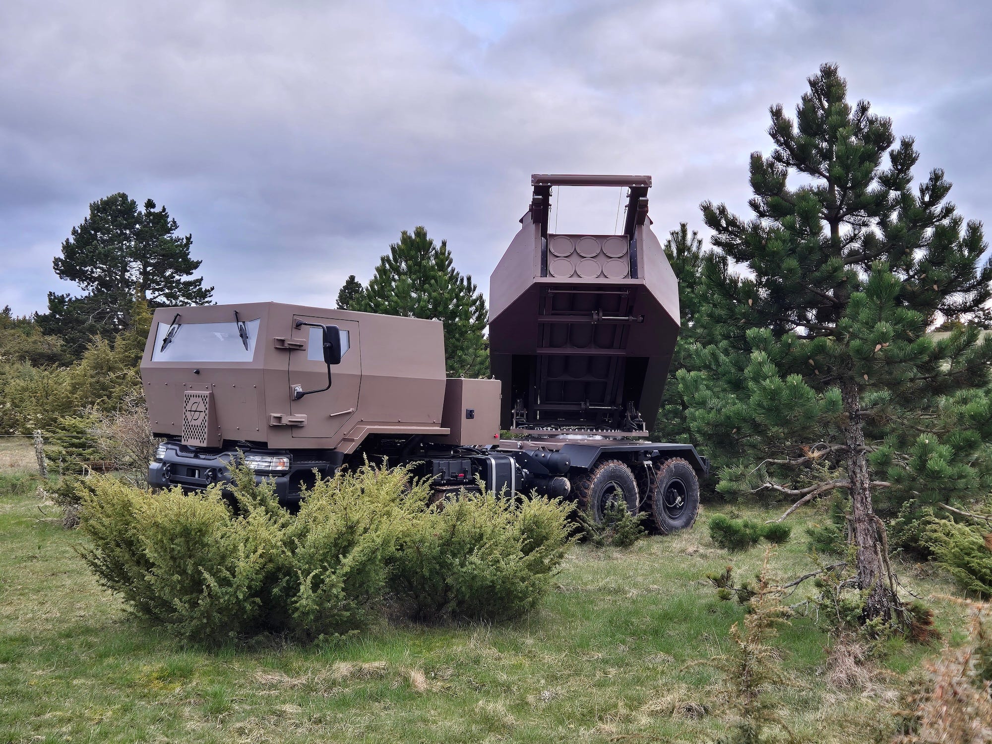A light brown rocket launder system on top of a truck on top of grass and among some trees