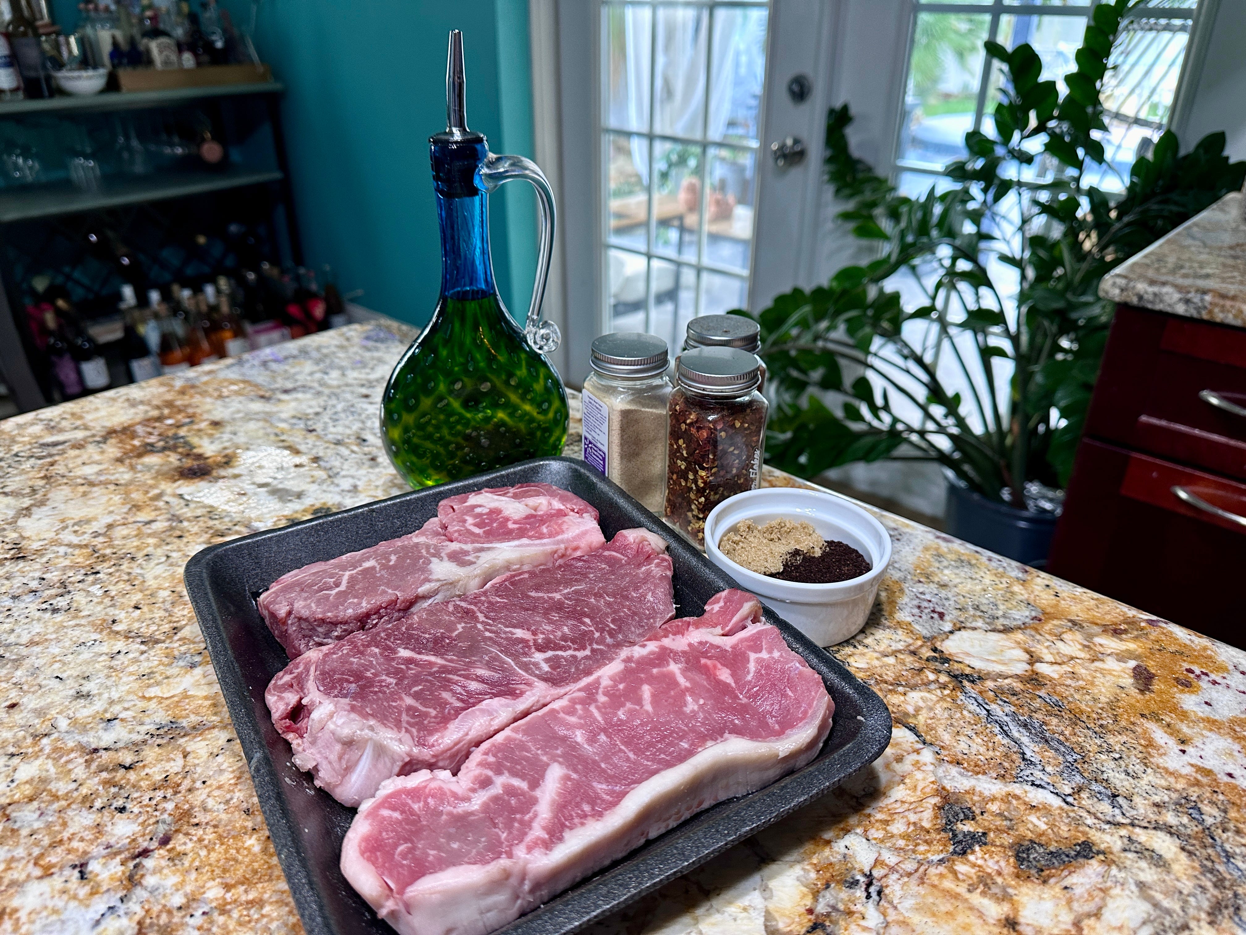 Three raw steaks in a baking pan, a container of olive oil, and various seasonings on a kitchen counter.