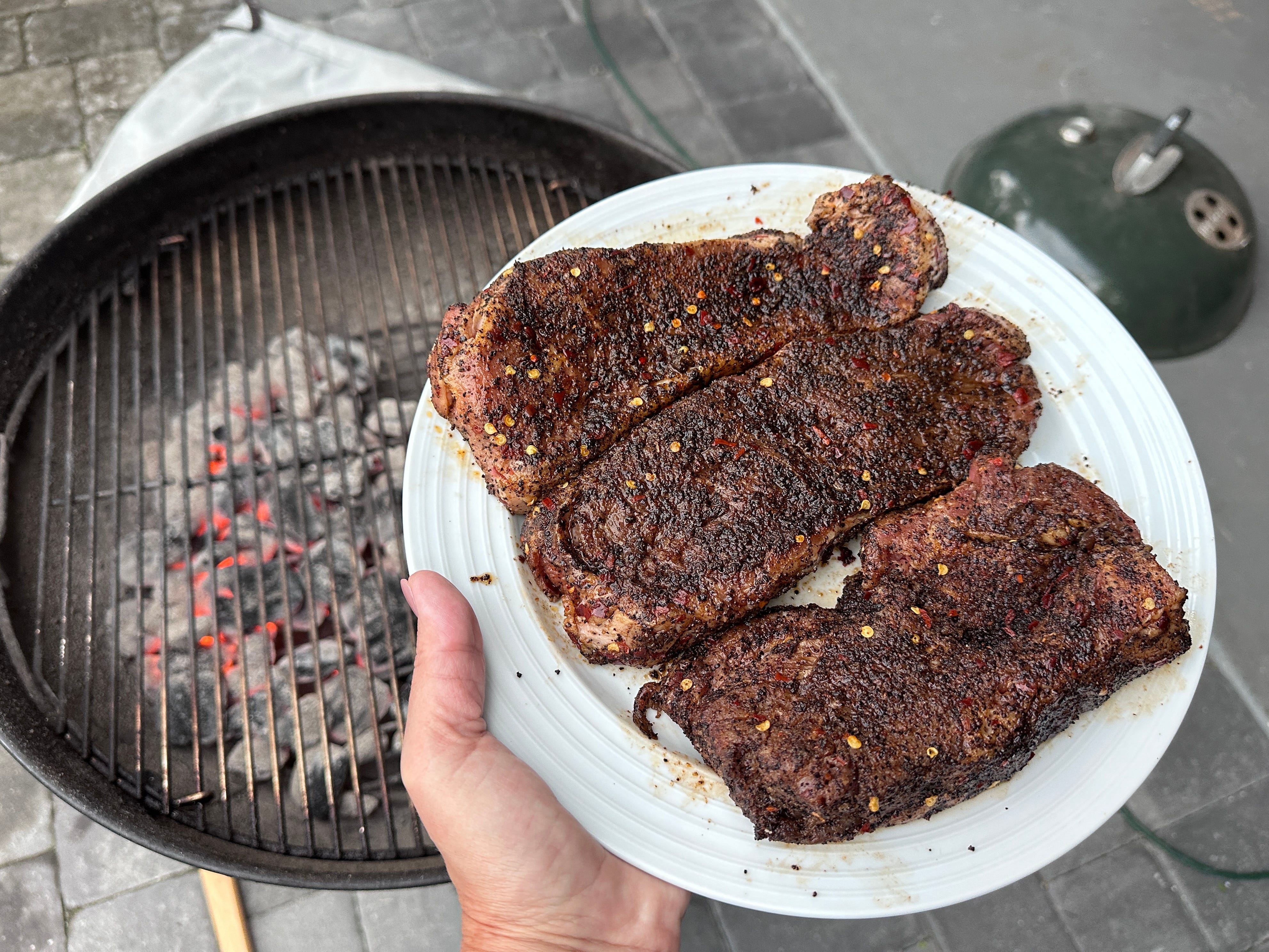 A hand holding a white plate with three pieces of steak over a charcoal grill.