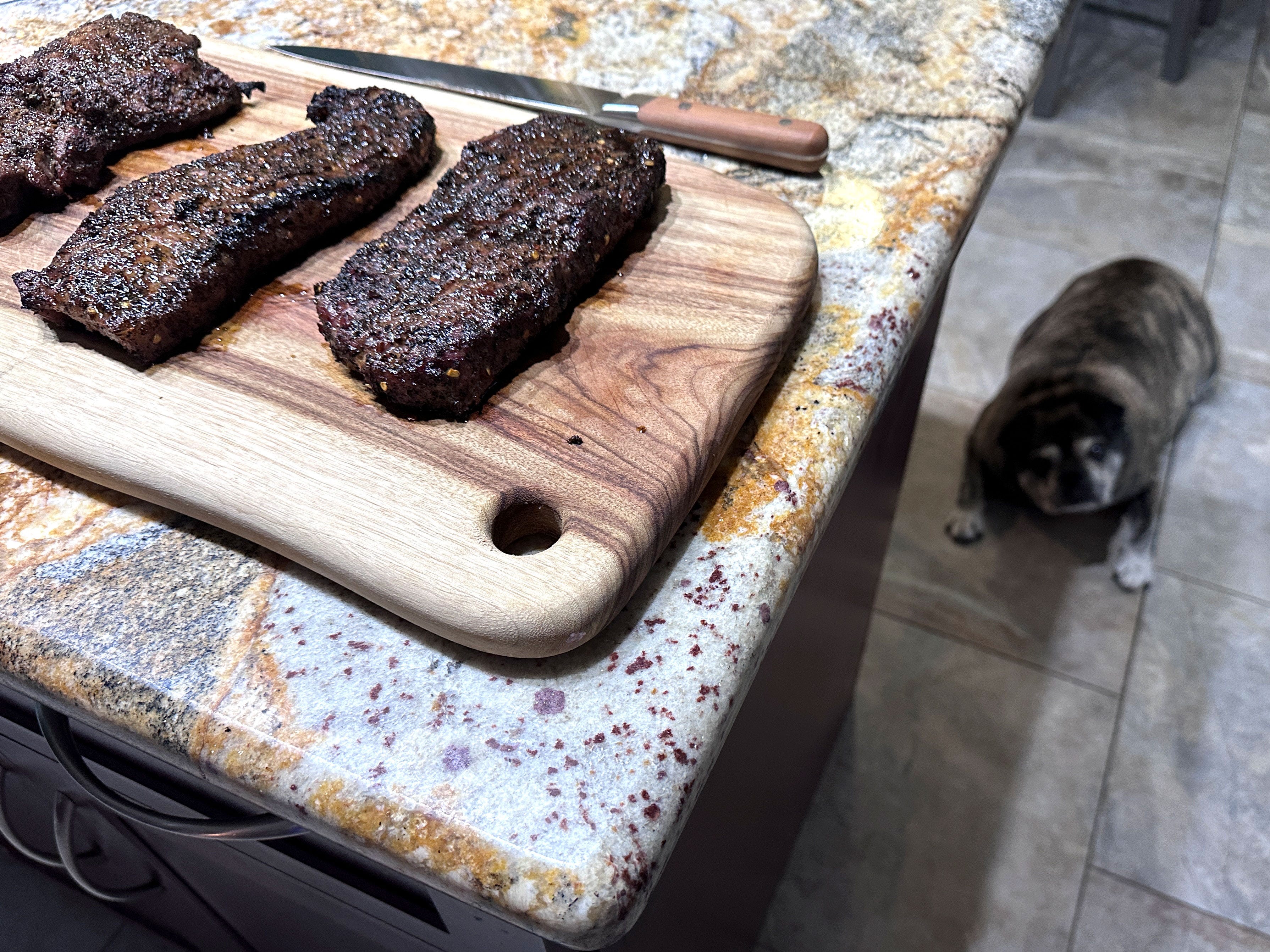 Three pieces of steak on a cutting board on a counter with a knife. A small dog lays next to the counter on the floor.