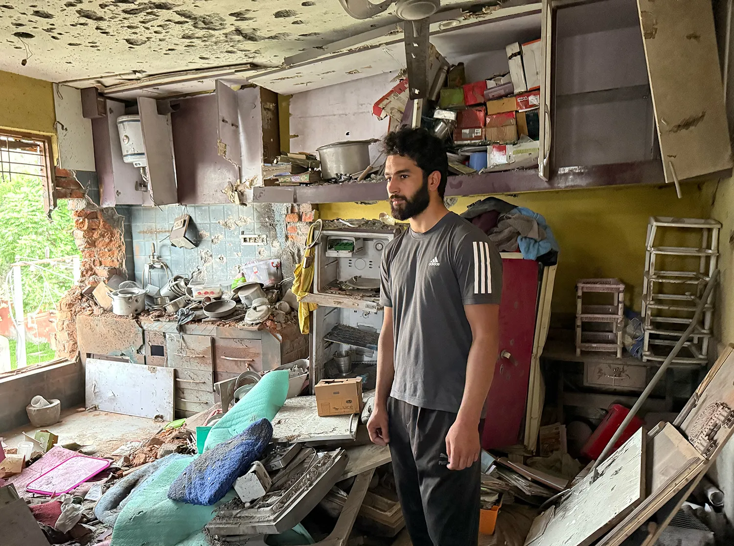 A man in a T-shirt looks stunned as he stands in a heavily damaged kitchen with a hole in the wall and debris all around him.
