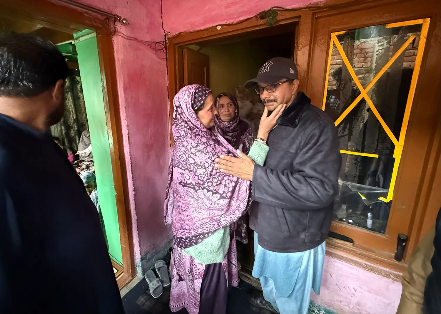 A man in a baseball cap stands in a doorway of a home with a sorrowful face as a woman in an patterned head scarf touches his face. Another woman is in the doorway and another man is at far left.