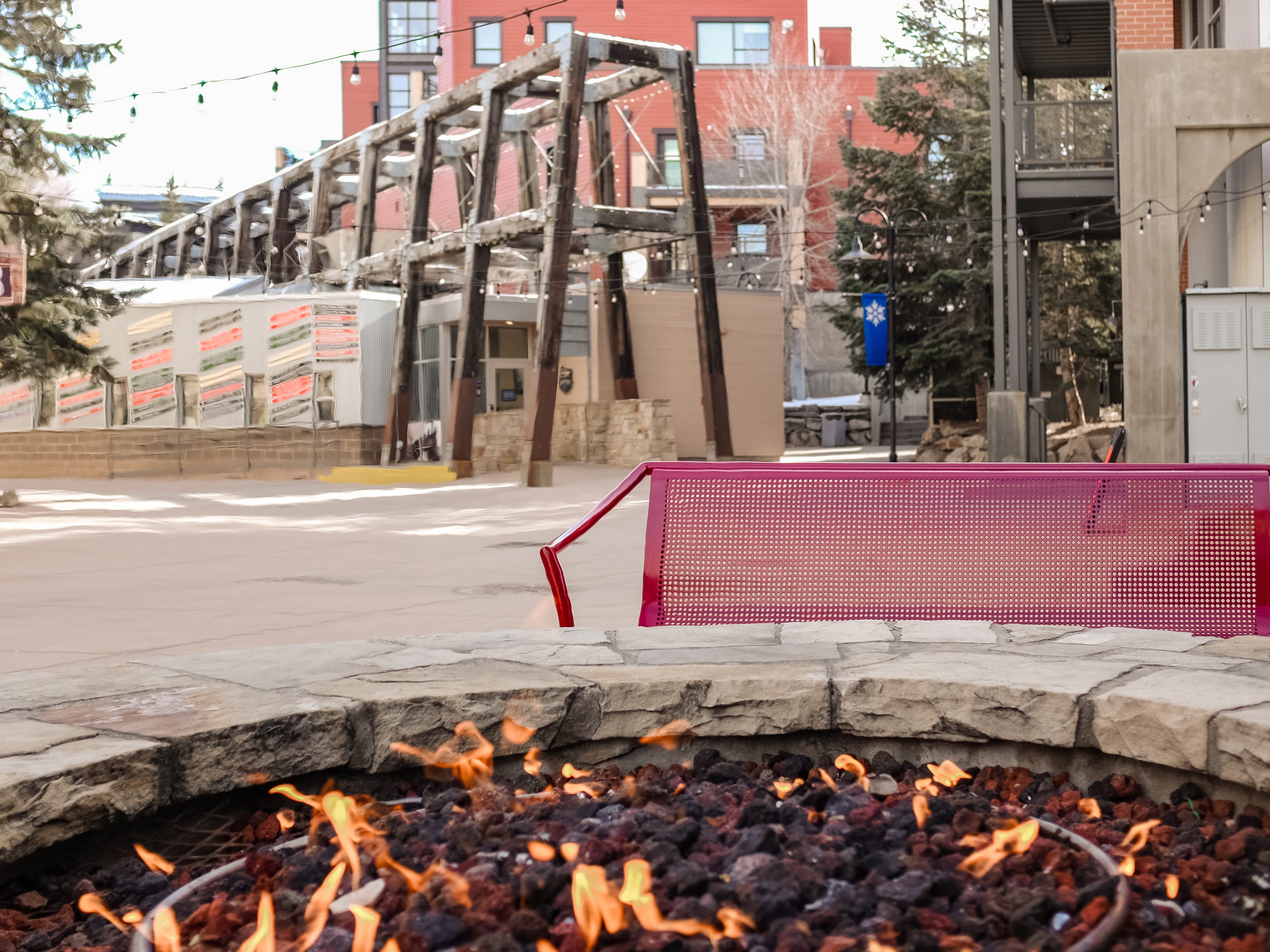 An outdoor fire pit in front of a red bench with trees and buildings in the background
