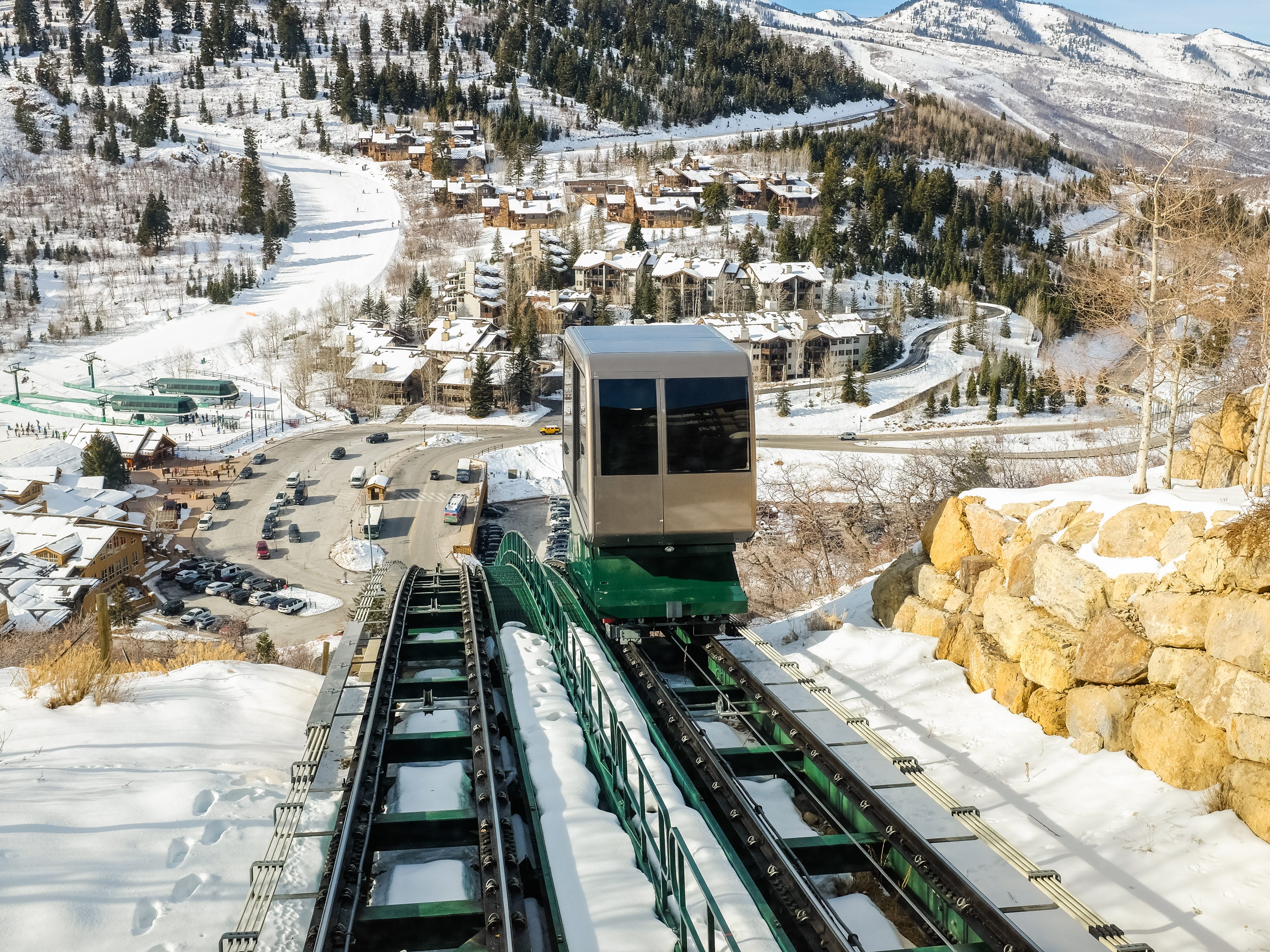 A funicular moves down a snowy mountain with a mountain topped with trees, housing, and snow in the background