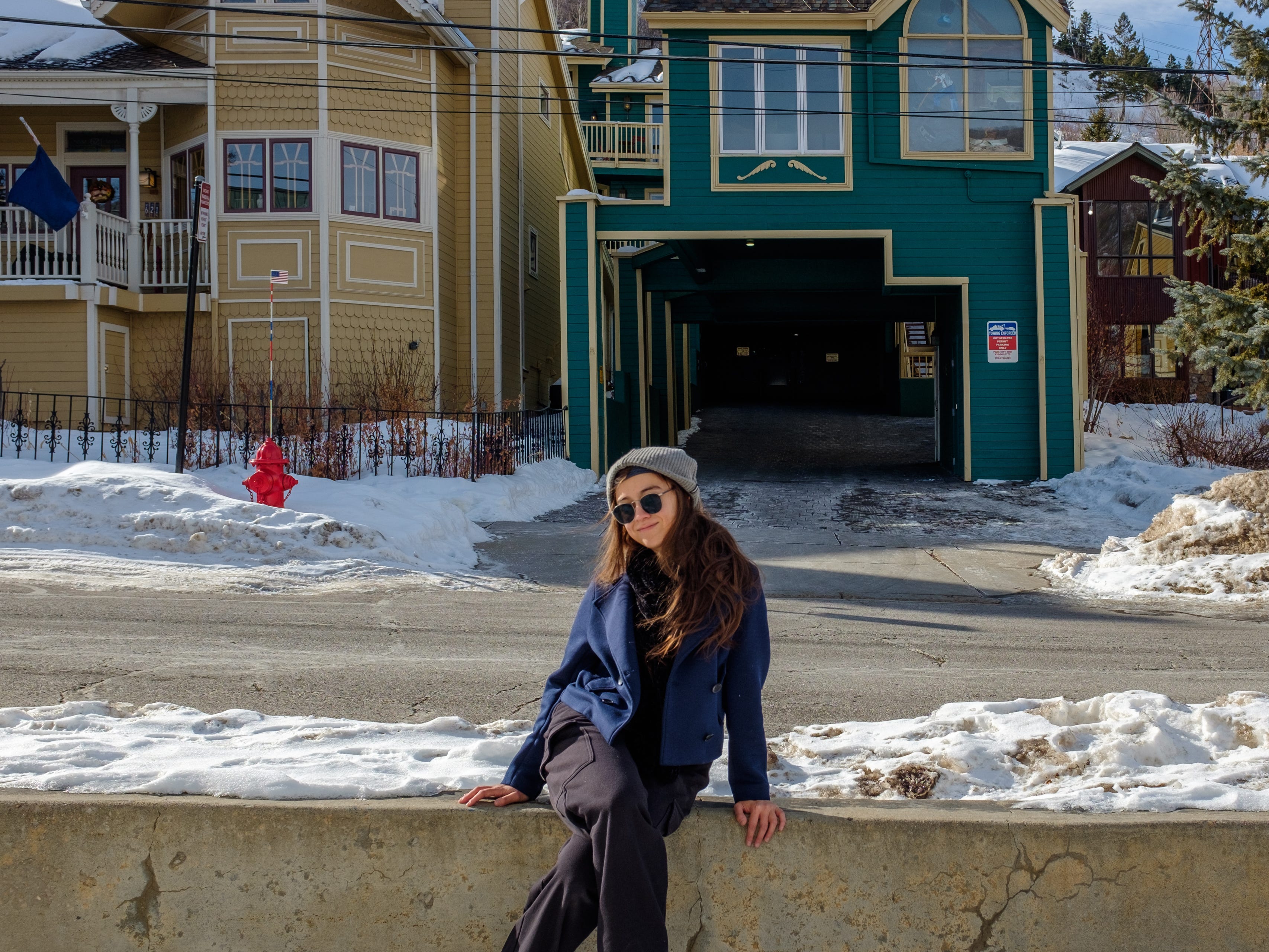 The author sits on a ledge next to a street with snow on the ground and houses in the beckground