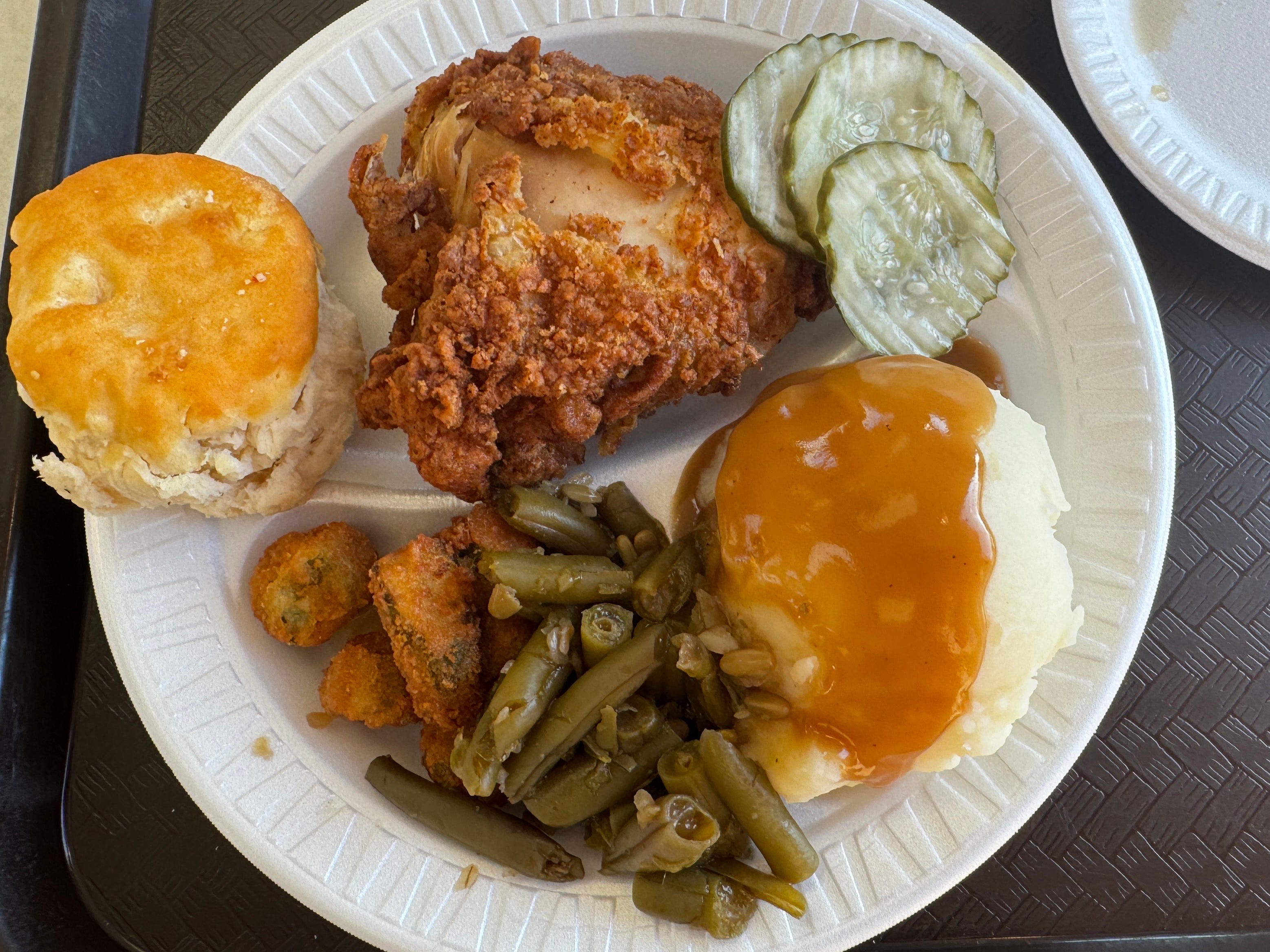 A biscuit, fried chicken, pickles, mashed potatoes with gravy, green beans, and fried okra on a styrofoam plate.