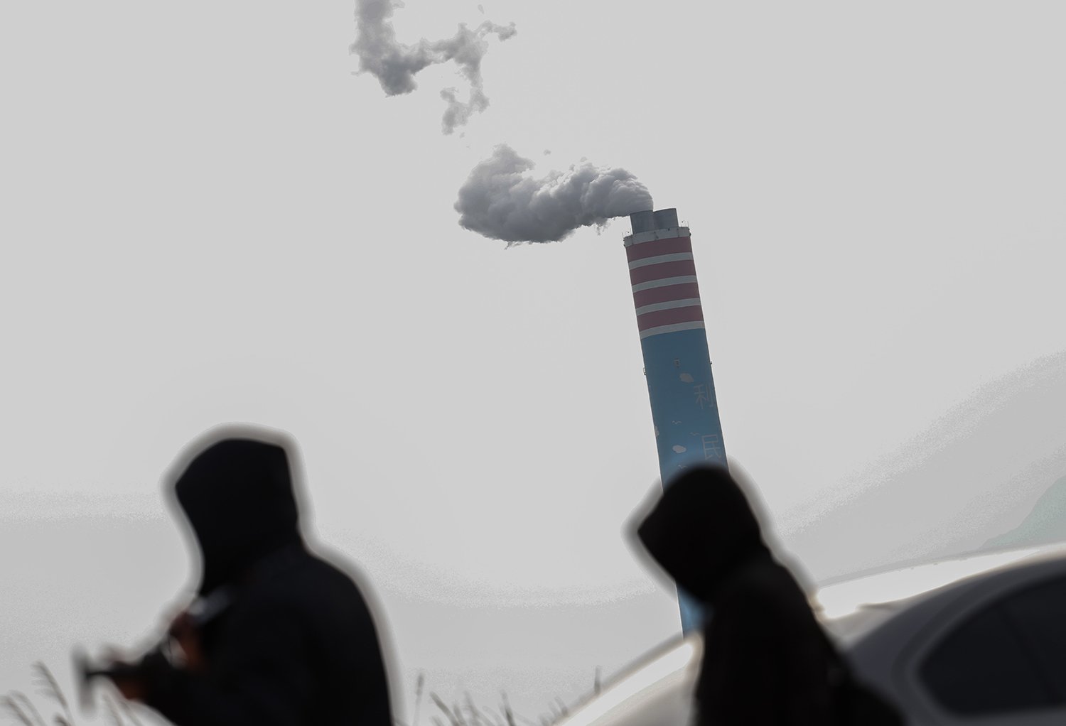 The blurry silhouettes of two figures are seen in front of a smokestack with a plume of smoke coming out of it.