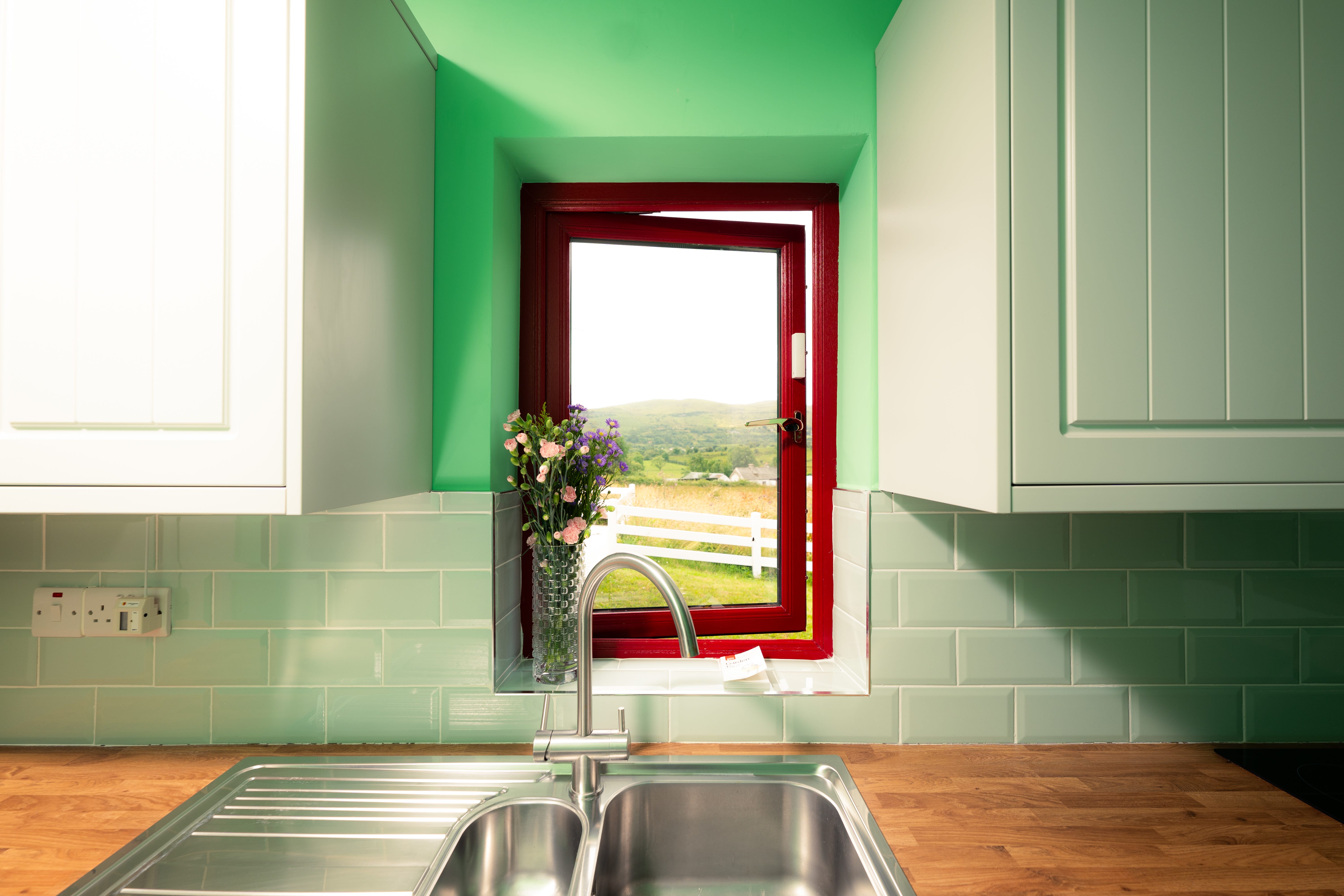 A window looking out on the Irish countryside silver-colored is set in a green wall above a kitchen sink and between two white cabinets