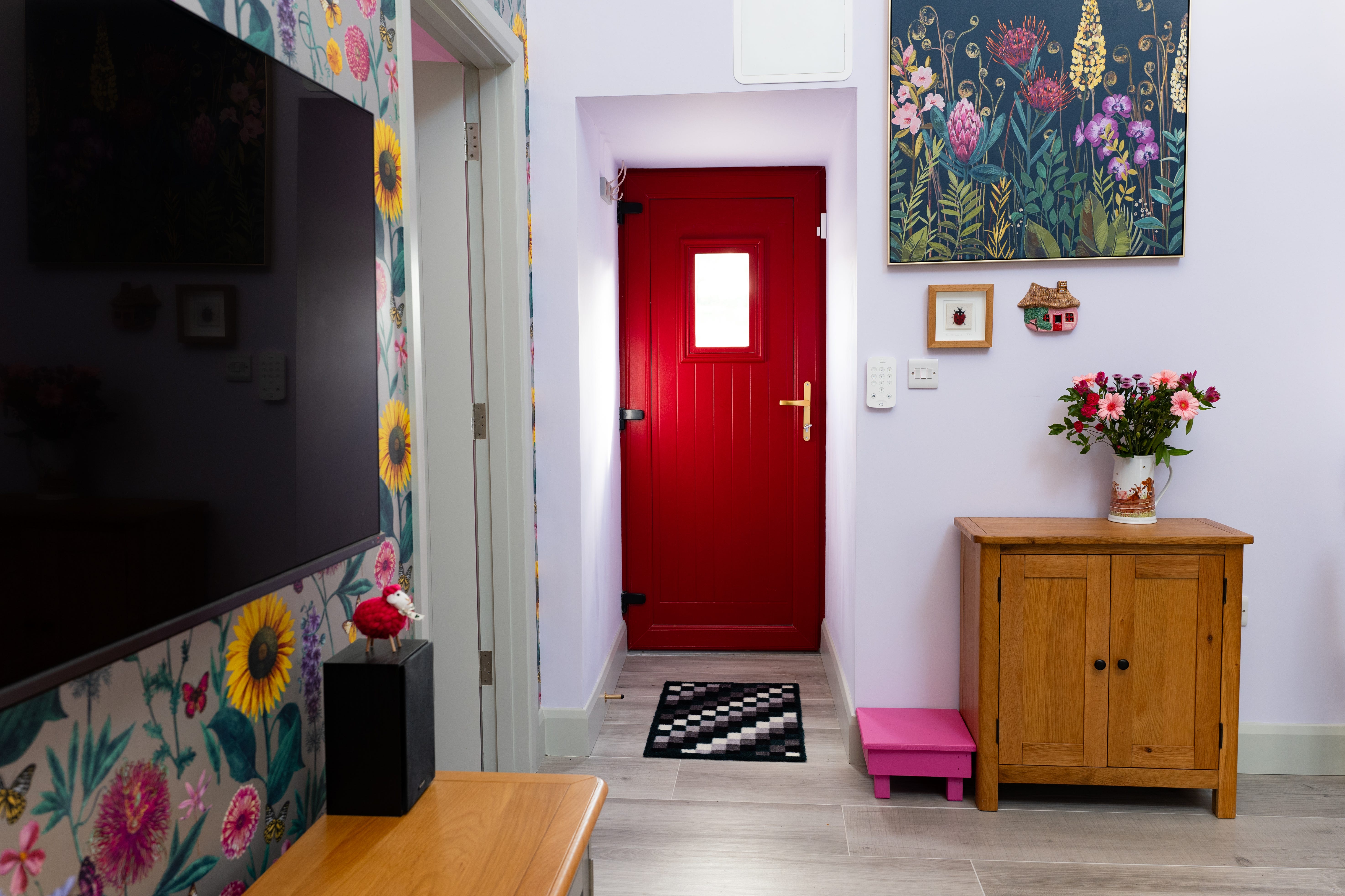 The back of a red front door and the home's entryway, with flowered wallpaper and art on the walls leading to it