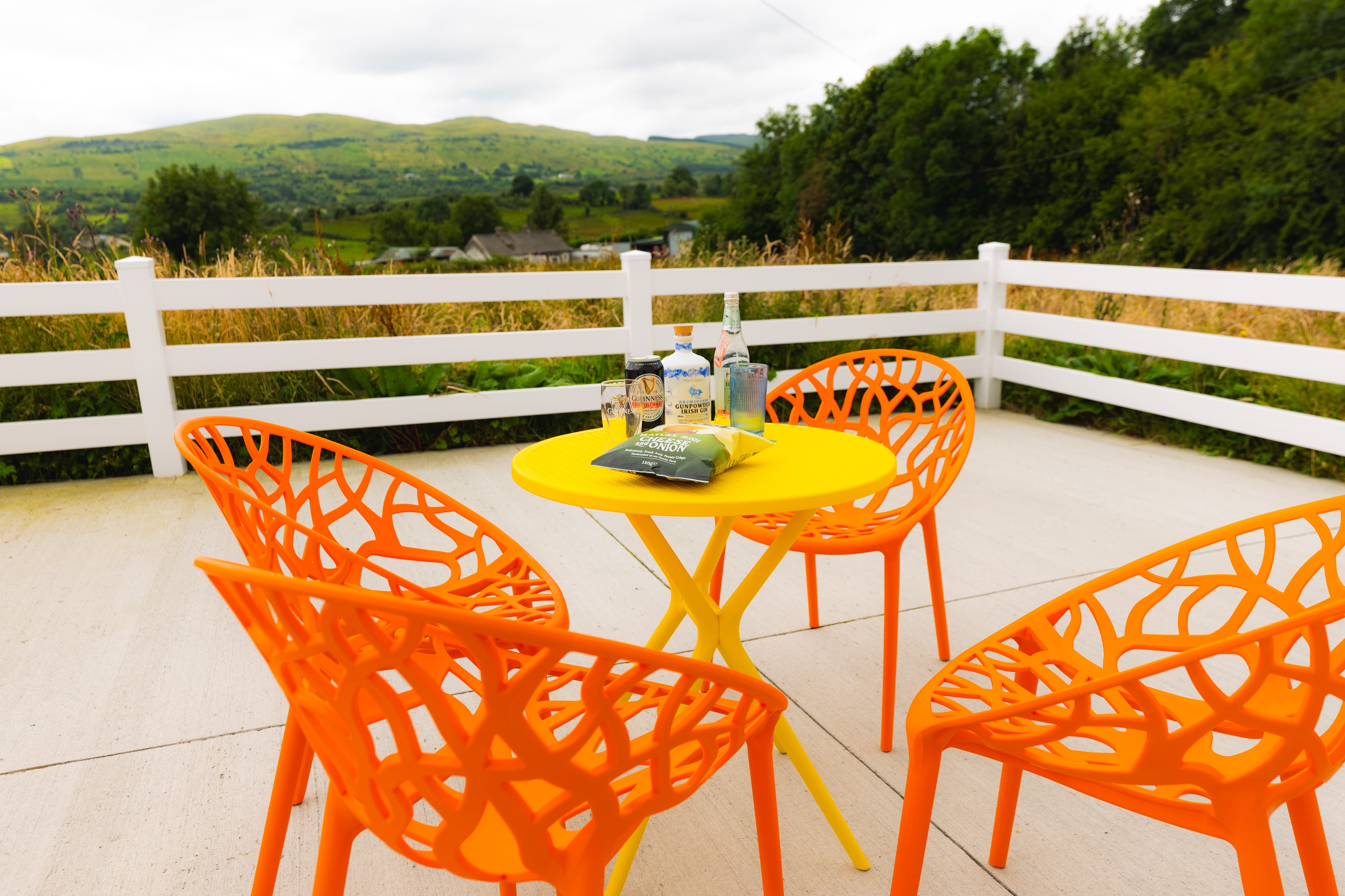 A yellow patio table with orange chairs sits on a patio surrounded by a white fence with the Irish countryside in the distance
