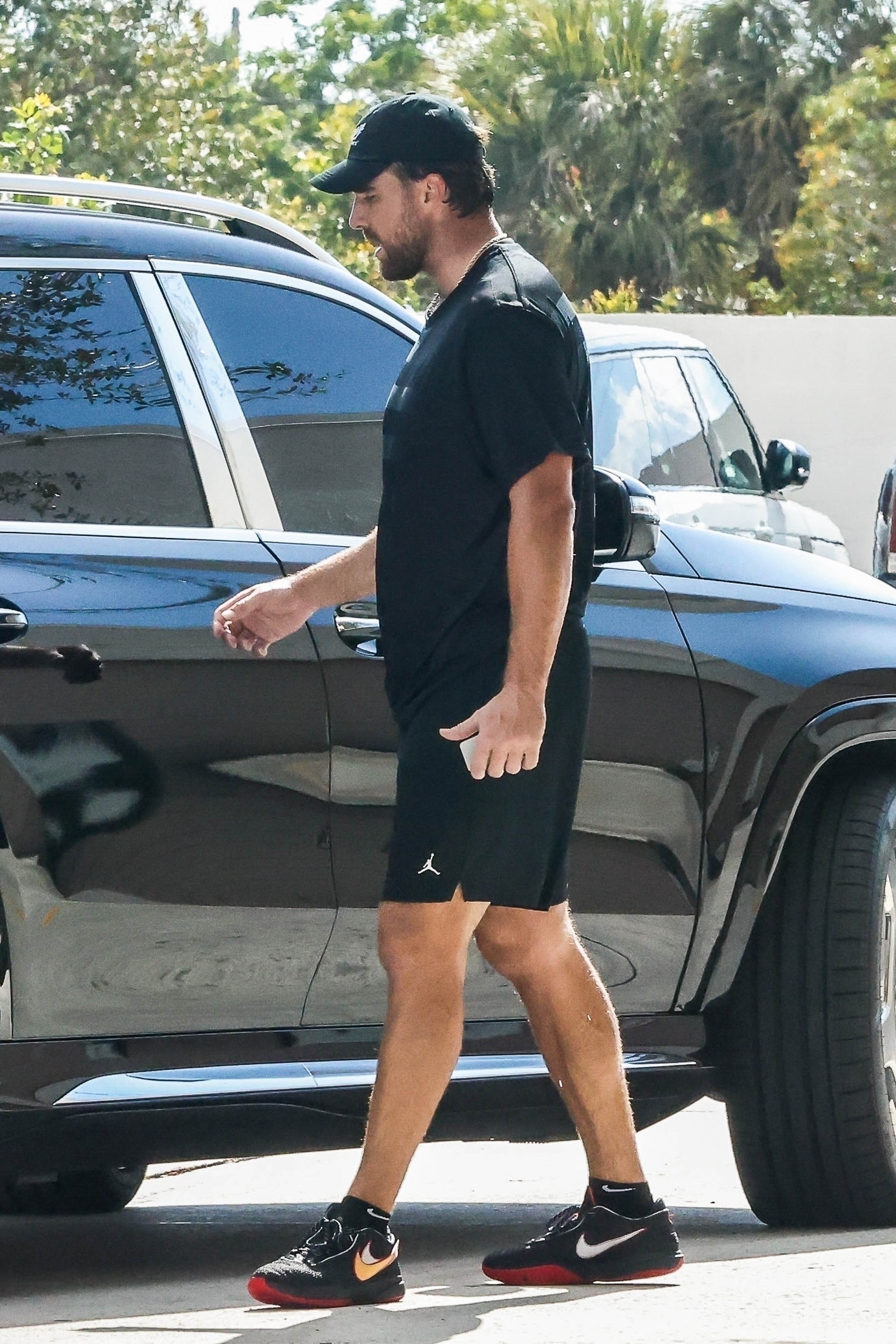 Travis Kelce by a vehicle in a black t shirt and black shorts. 