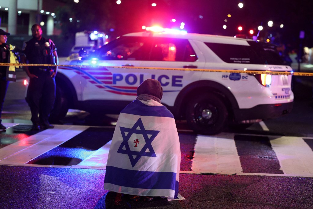 A man, with an Israeli flag with a cross in the center, kneels next to emergency personnel working at the site where, according to the U.S. Homeland Security Secretary, two Israeli embassy staff were shot dead near the Capital Jewish Museum in Washington, D.C., U.S. May 21, 2025.