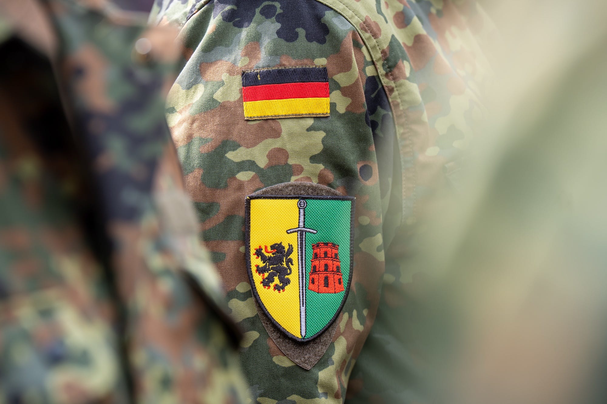 A patch on the arm of a camouflage jacket that on one side shows a lion against a yellow background and on the other shows a red tower against a green background, with a sword running down between the two sides