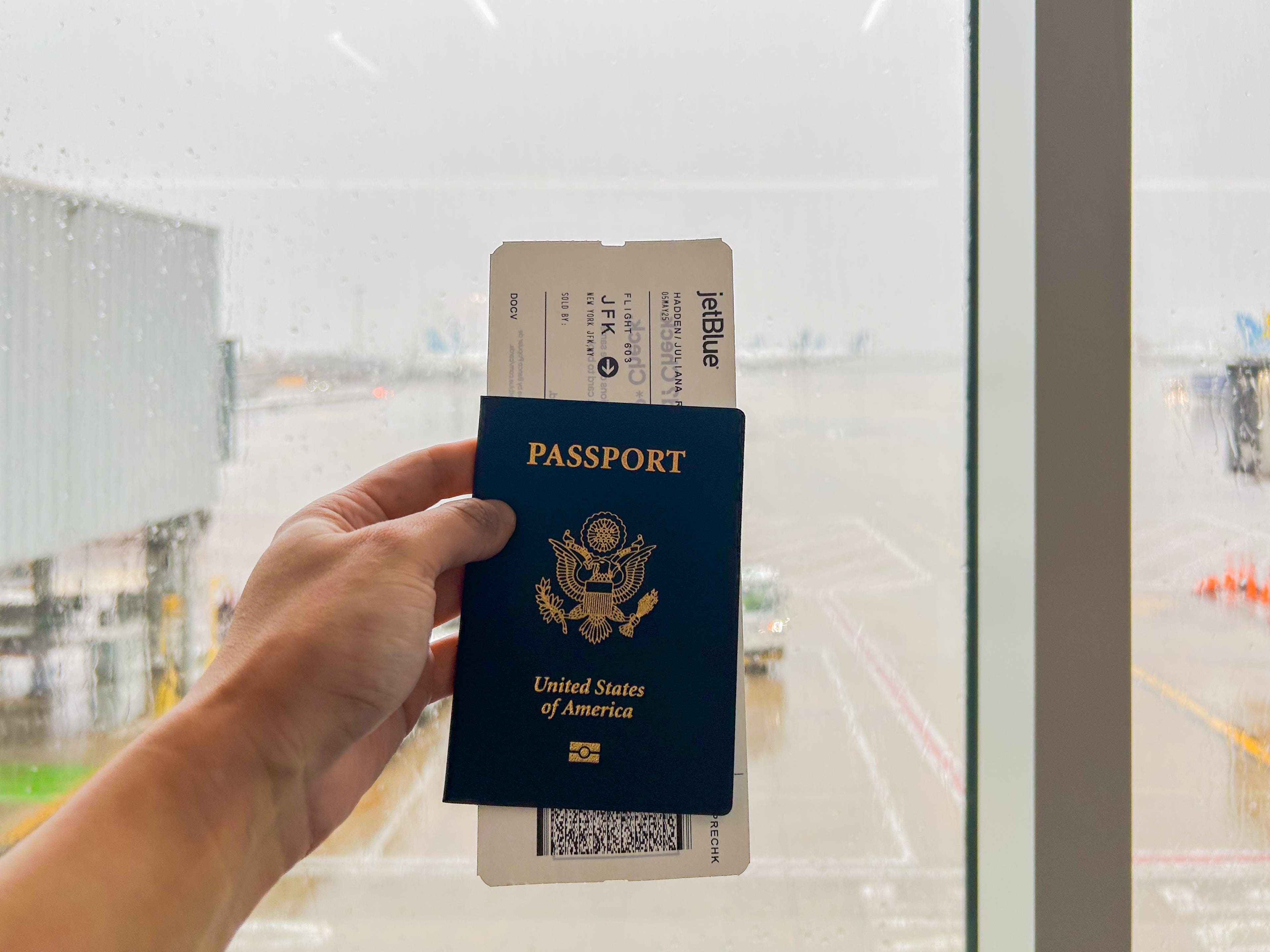 A hand holds a passport with a boarding pass inside in front of a window at an airport