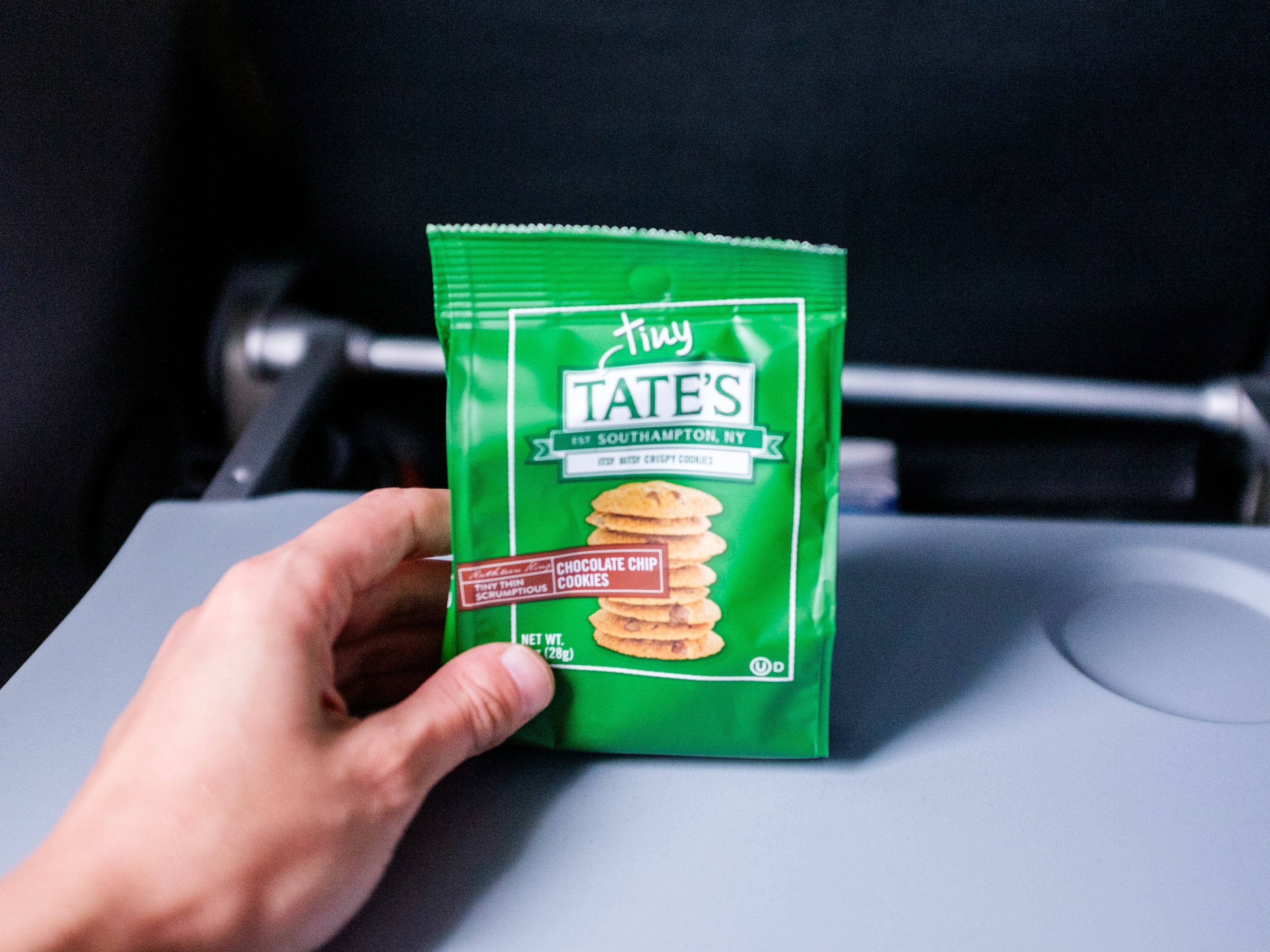 A hand holds a tiny green package of Tate's chocolate chip cookies on a tray table on a plane
