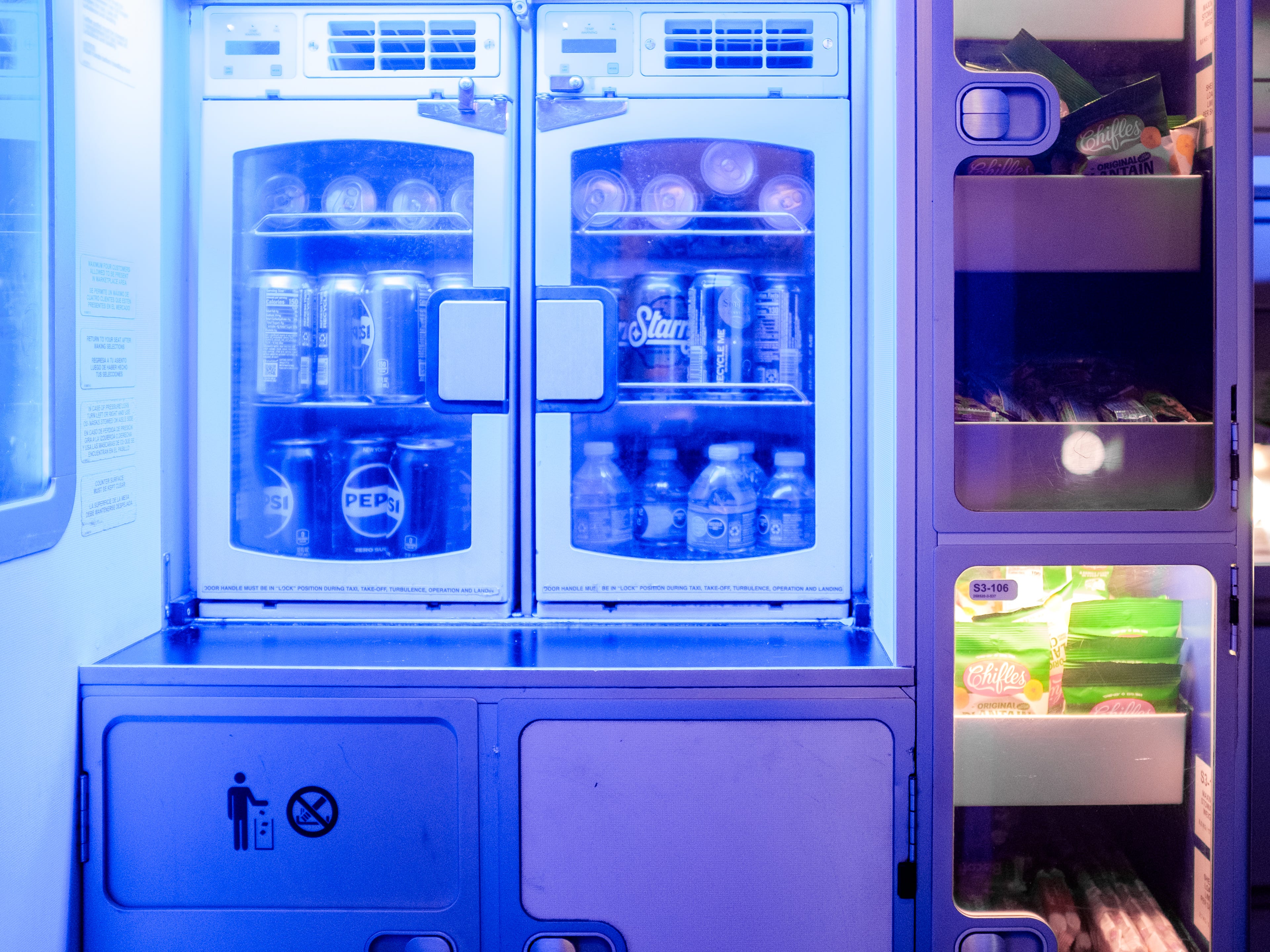 A JetBlue pantry with a blue glow has a mini fridge stocked with water and sodas on the left and cabinets of snacks on the right