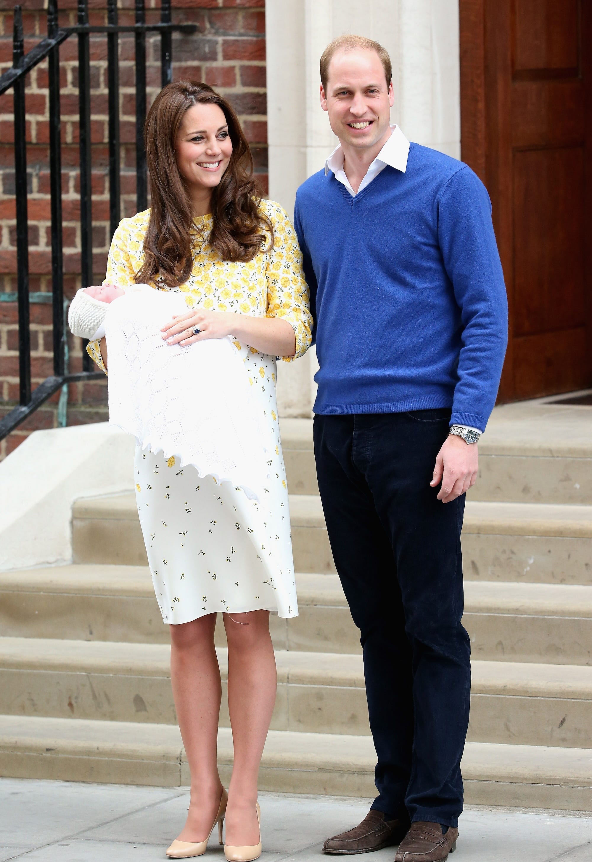 Kate Middleton, dressed in a yellow and white dress, and Prince William, dressed in a royal blue long-sleeved shirt and jeans, pose with newborn Princess Charlotte in front of hospital steps.