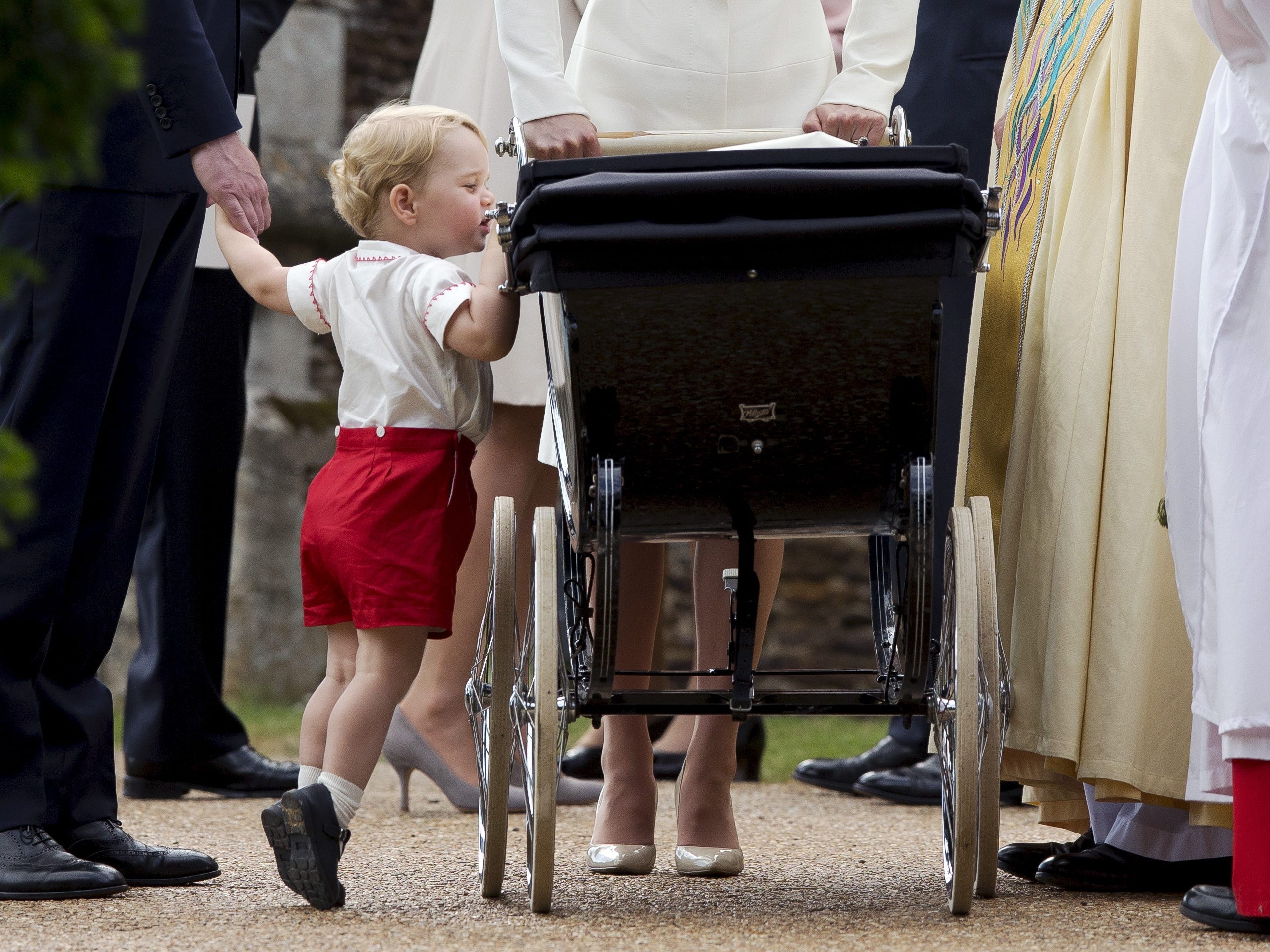 Prince George, dressed in a white shirt, red shorts, and black shoes, peers into a black pram holding his sister, Princess Charlotte.