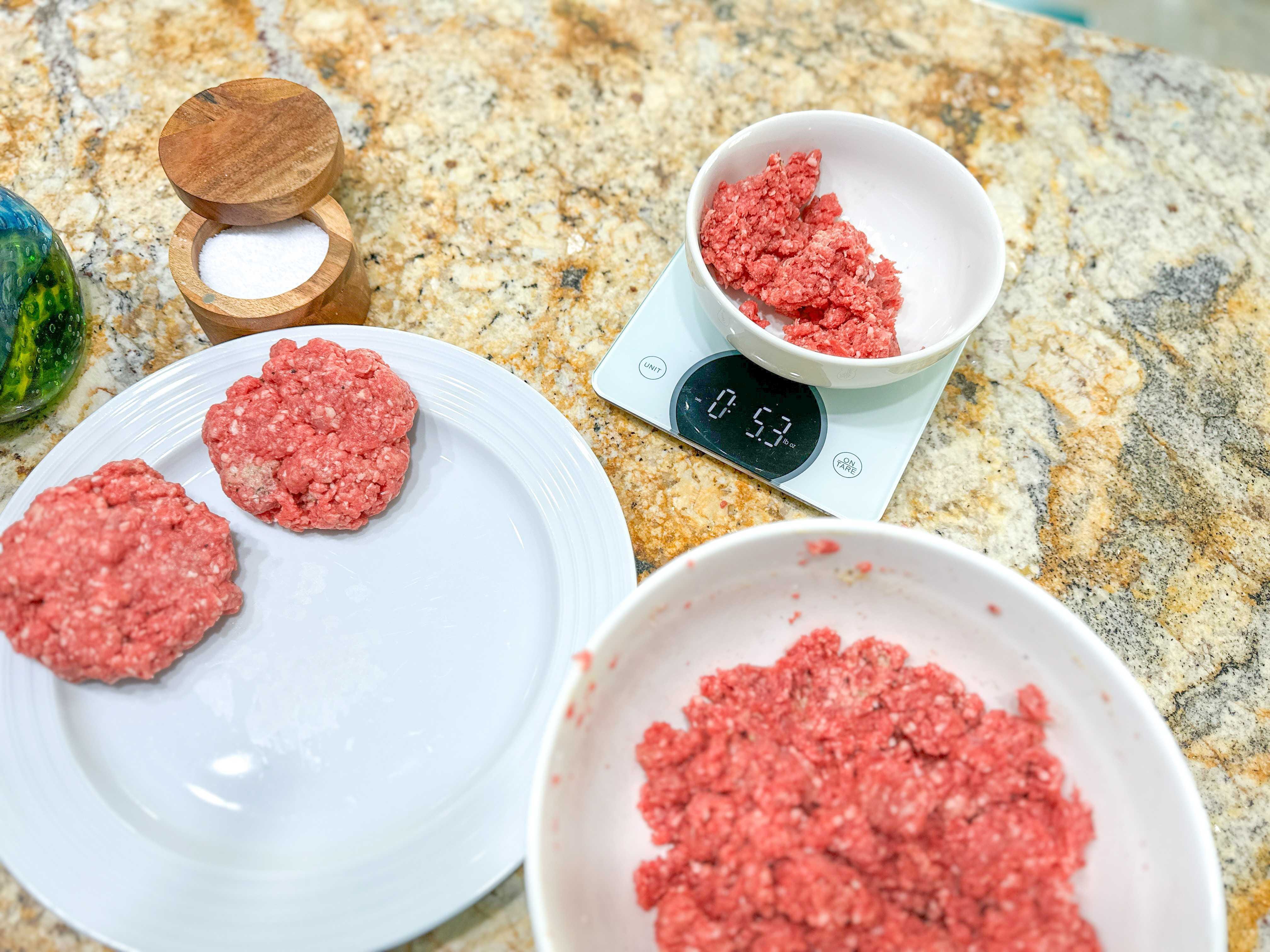 A large bowl of ground beef sitting next to a white plate with two burger patties and a smaller white bowl of ground beef sitting on a food scale