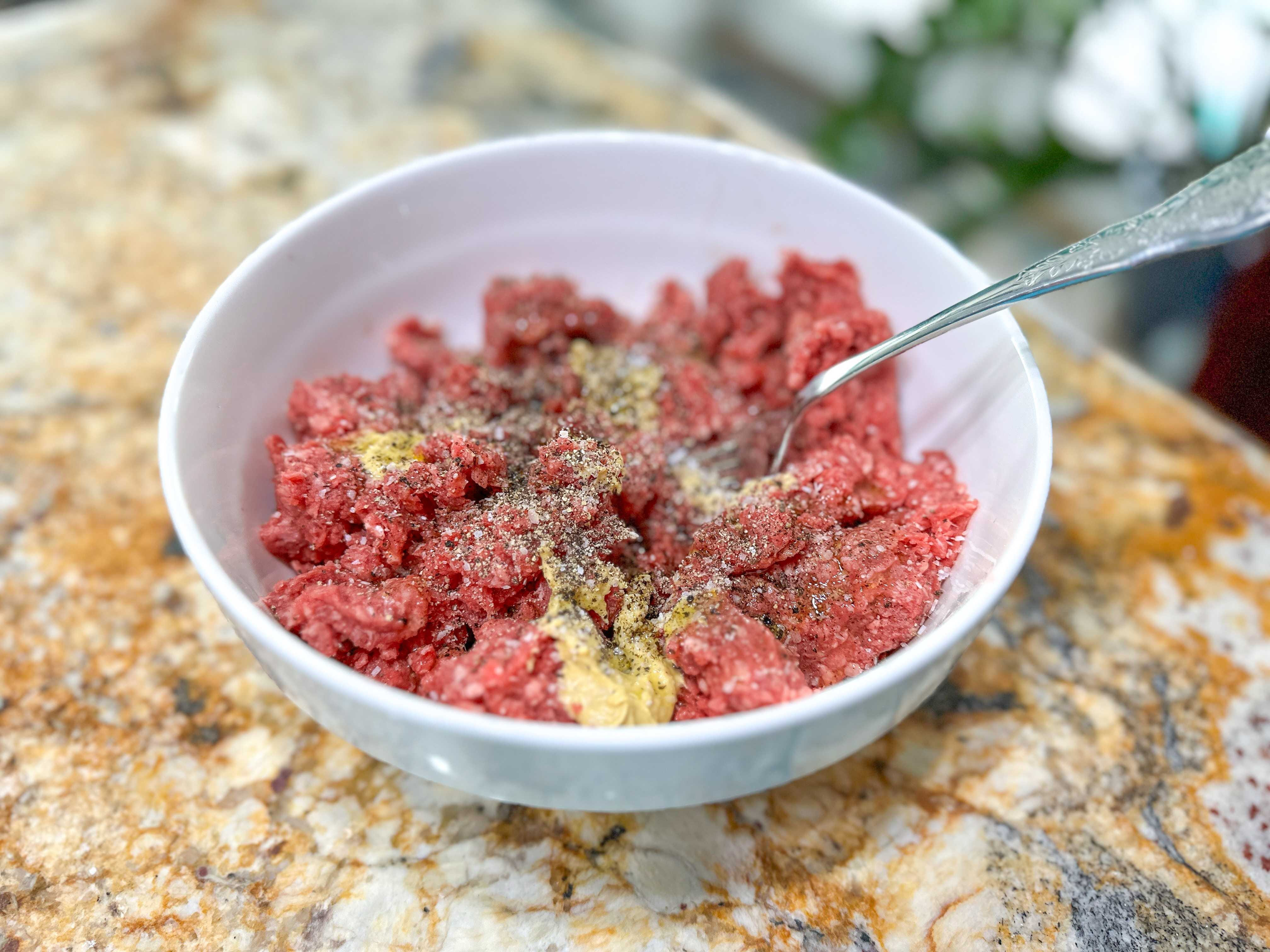 A white bowl of ground beef with salt, pepper, mustard, and a fork sticking out of the mixture