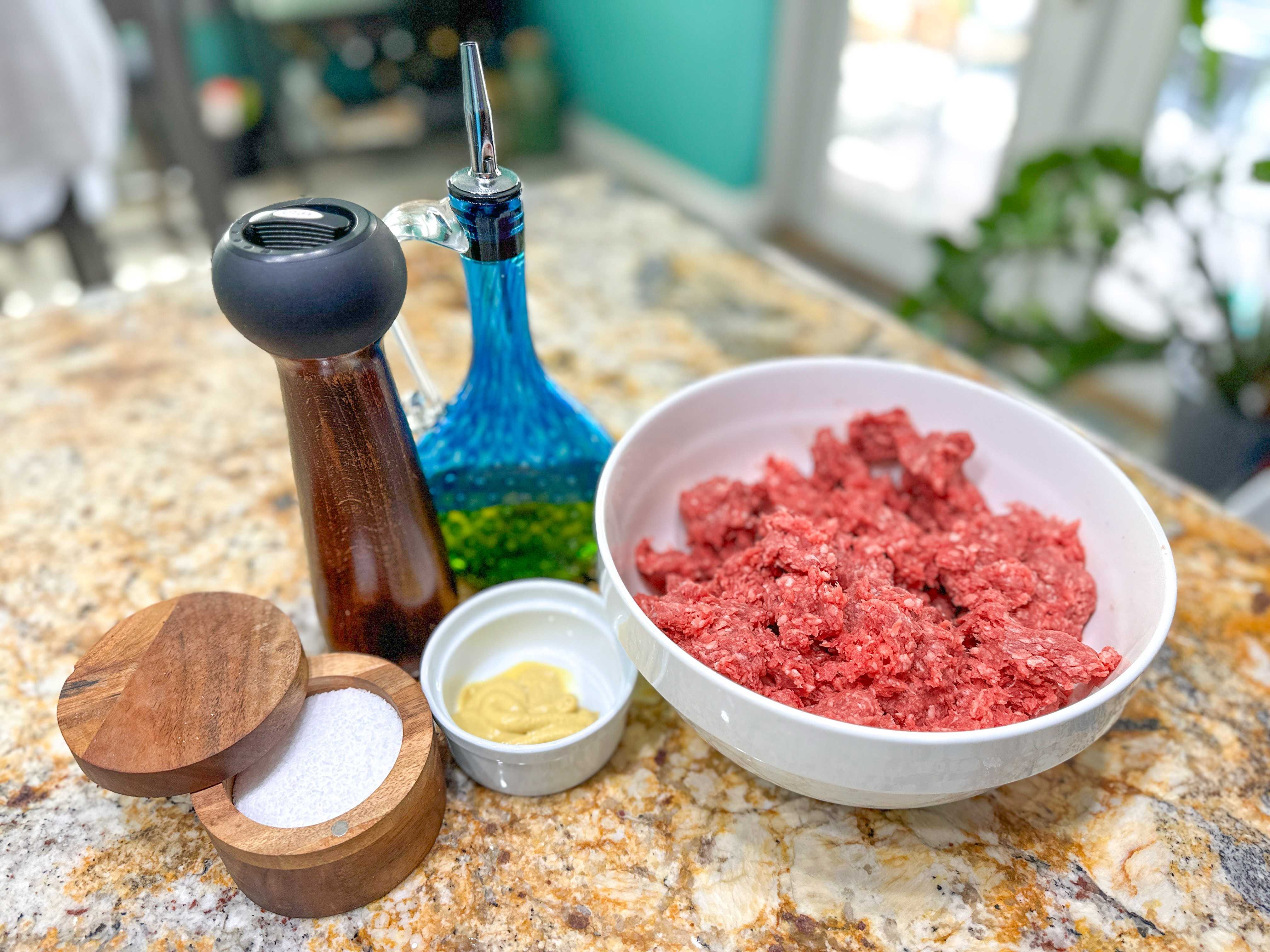 A white bowl filled with ground beef, a pepper grinder, a small white bowl of mustard, a brown bowl of salt, and a blue olive oil dispenser
