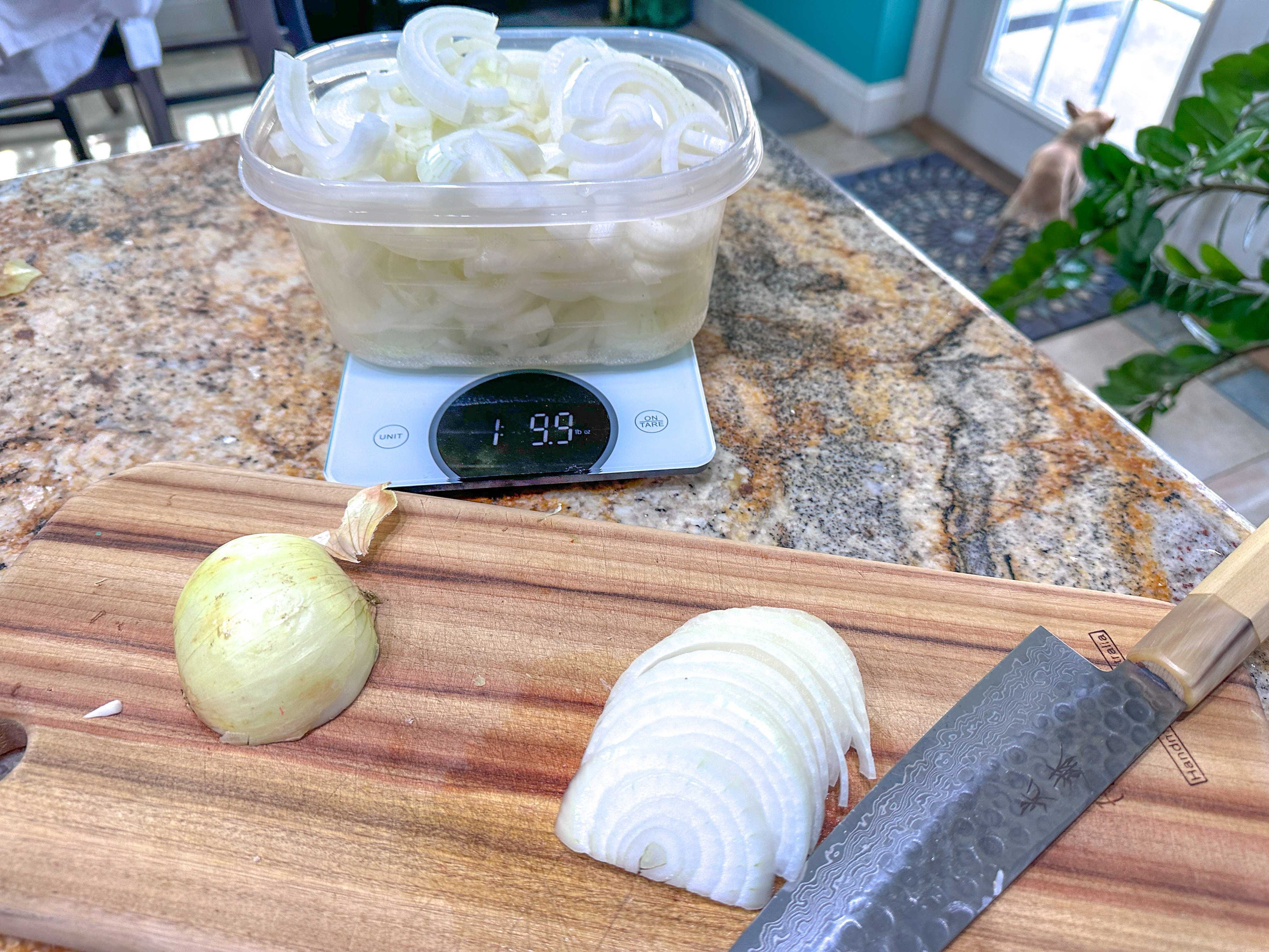 A wooden cutting board with a half of an onion, some onion slices, and a knife sitting in front of a container of onion slices on a food scale