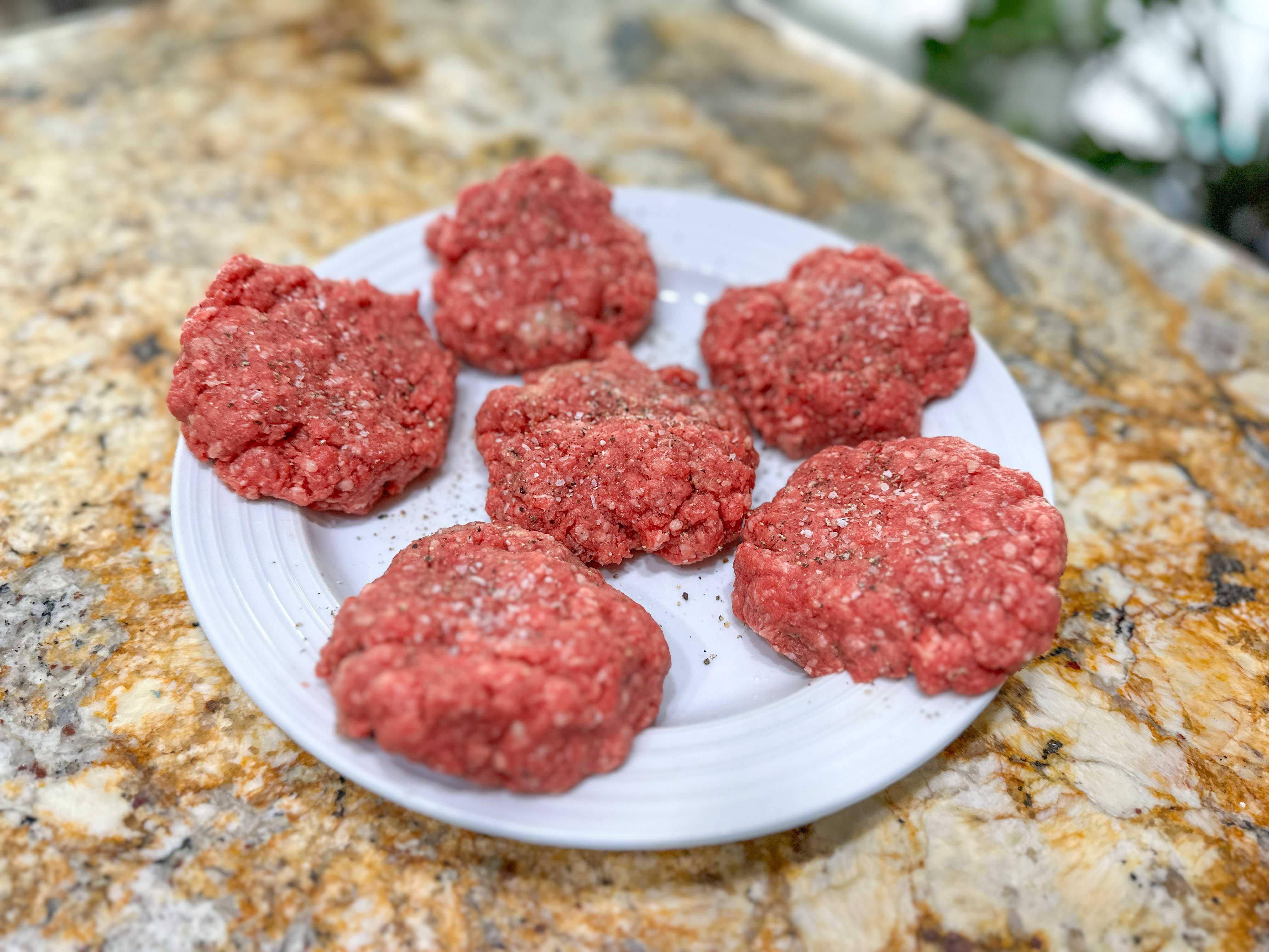A white plate of six raw burger patties sprinkled with salt and pepper