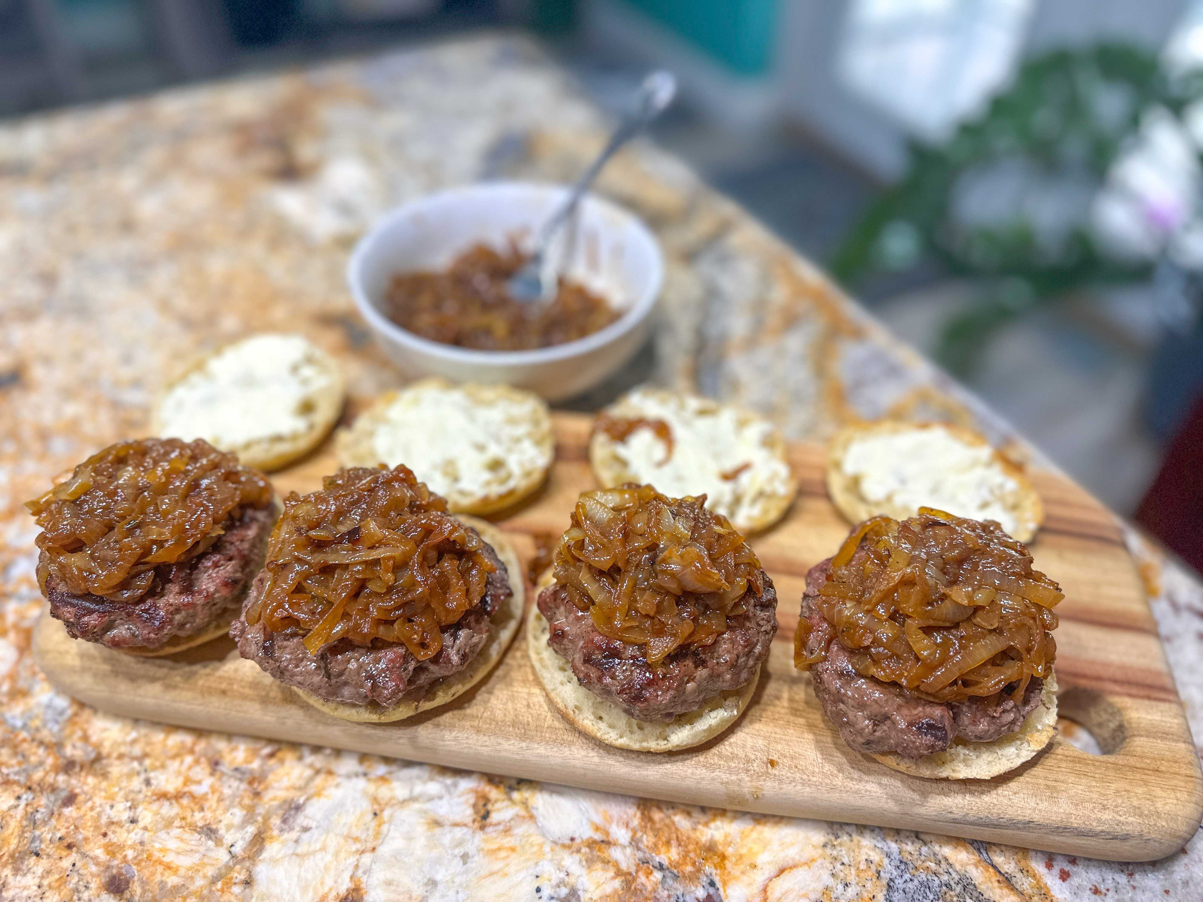 A wooden board holding four burger patties sitting on top of English muffin buns with caramelized onion on top of them. English muffin tops with a white spread sit on the board behind them.