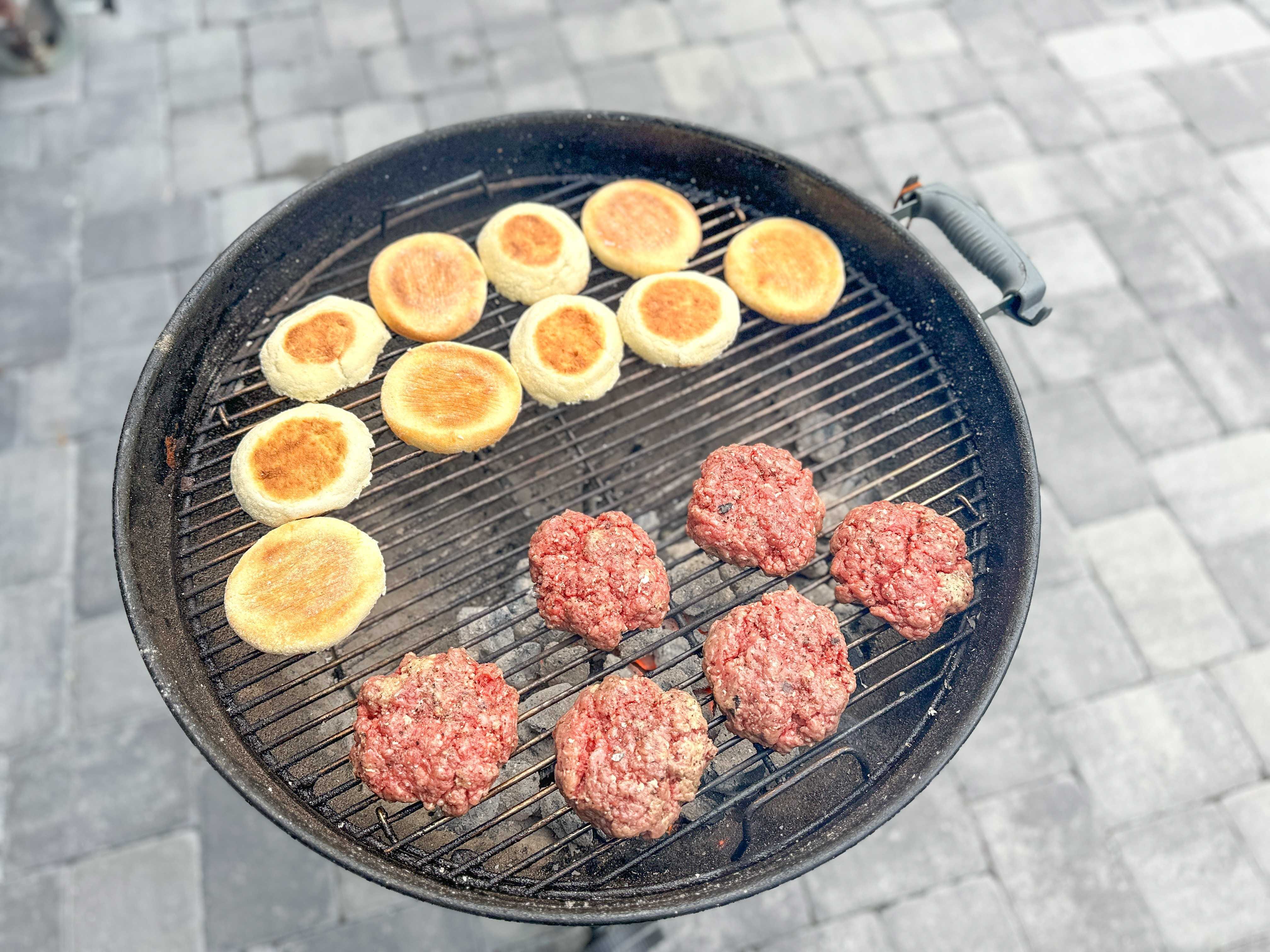 Six raw burger patties sitting on a grill next to several English muffin buns