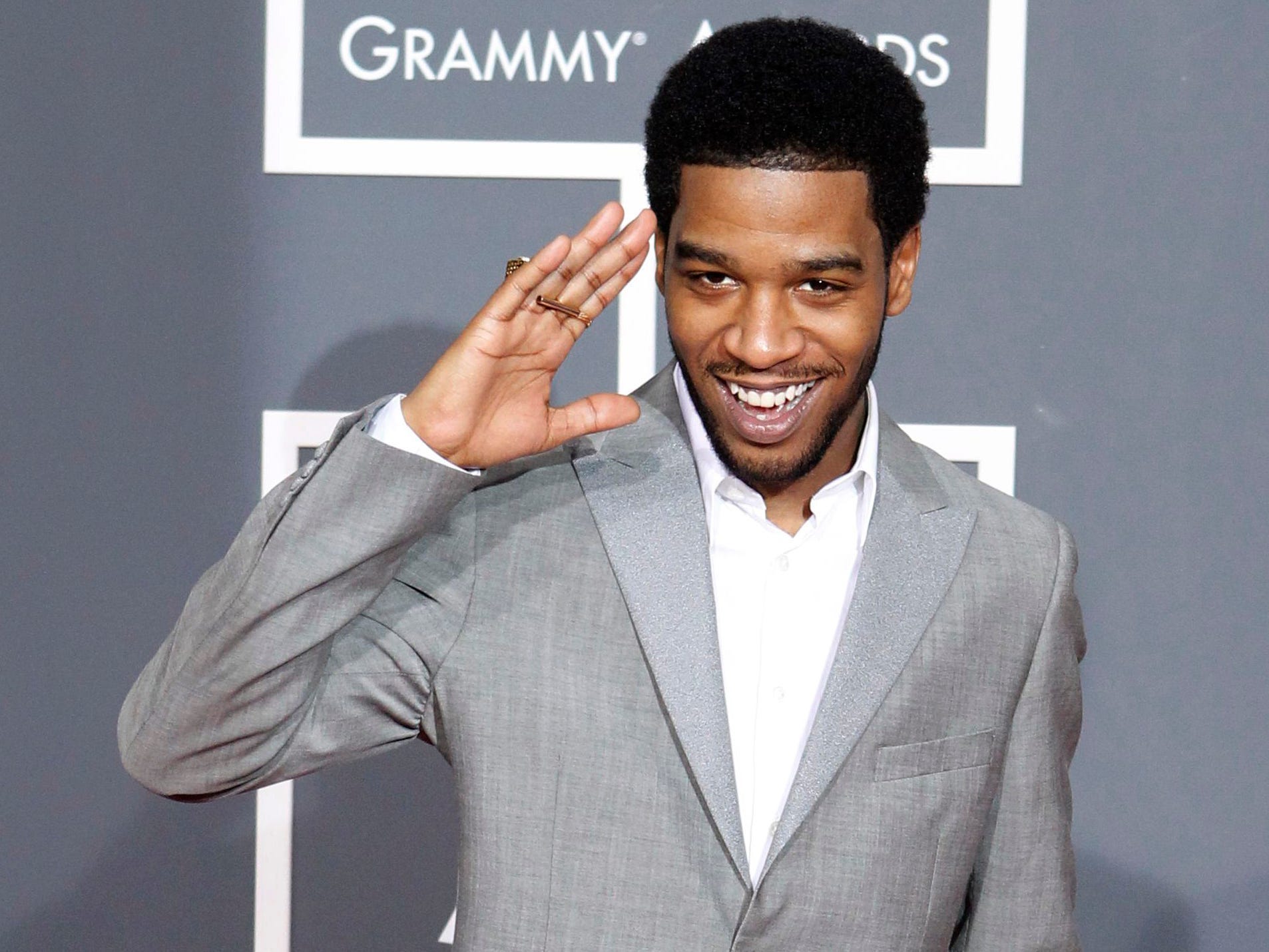Kid Cudi in a grey suit waving with his right hand and his left hand in his trouser pockets.