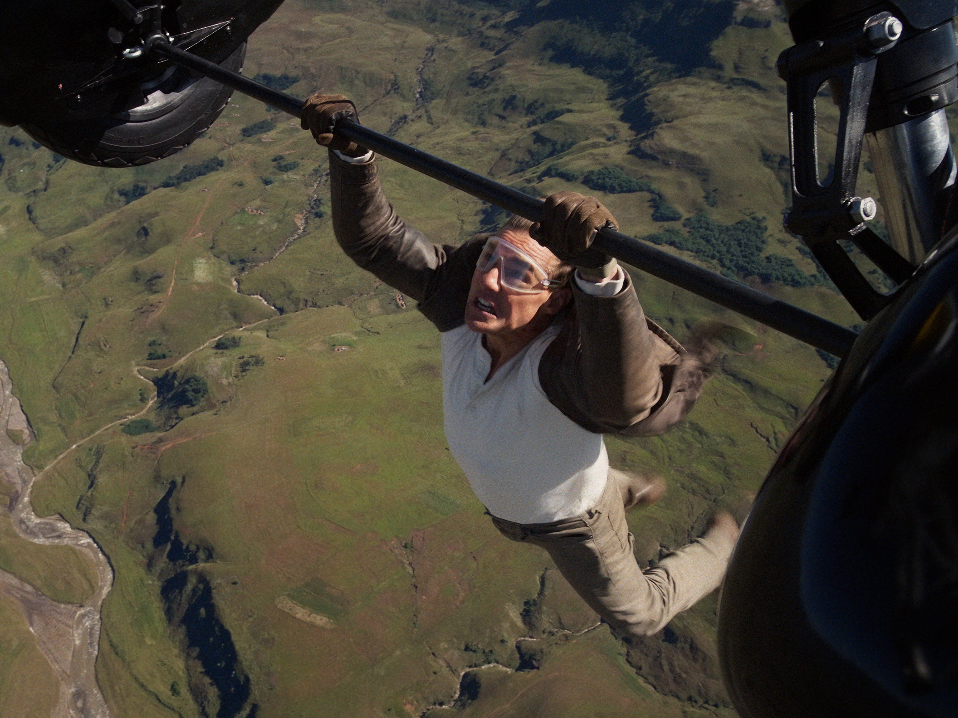 A man wearing a brown leather jacket. a white long-sleeved shirt, brown pants, and shoes. He's clinging onto a set of aircraft wheels while flying without a harness. He's also wearing a pair of goggles.