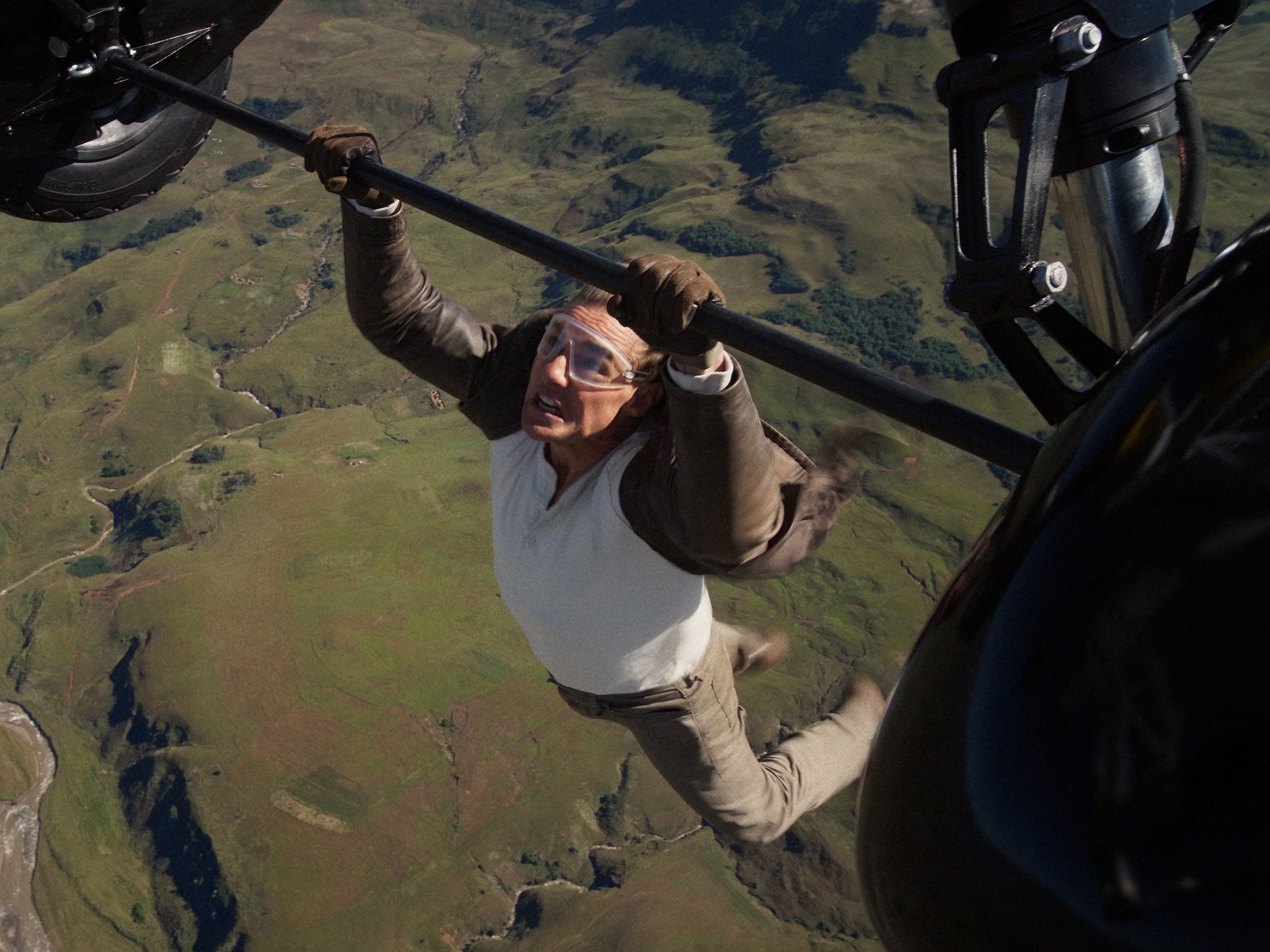 A man wearing a brown leather jacket. a white long-sleeved shirt, brown pants, and shoes. He's clinging onto a set of aircraft wheels while flying without a harness. He's also wearing a pair of goggles.