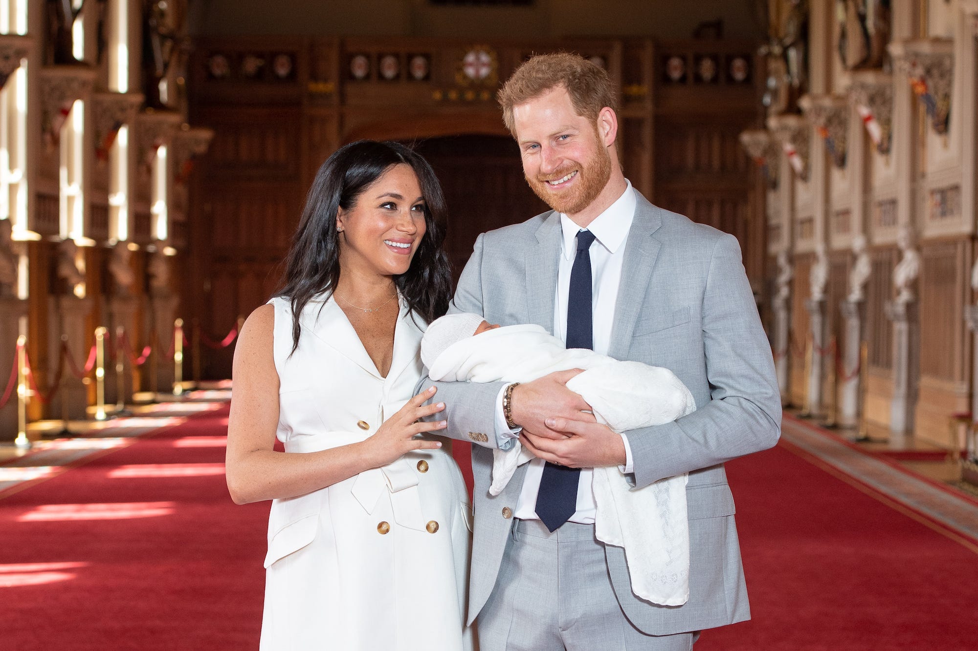 Meghan Markle, dressed in a white dress, posed with Prince Harry, dressed in a gray suit, and their son Archie, wrapped in a white blanket.