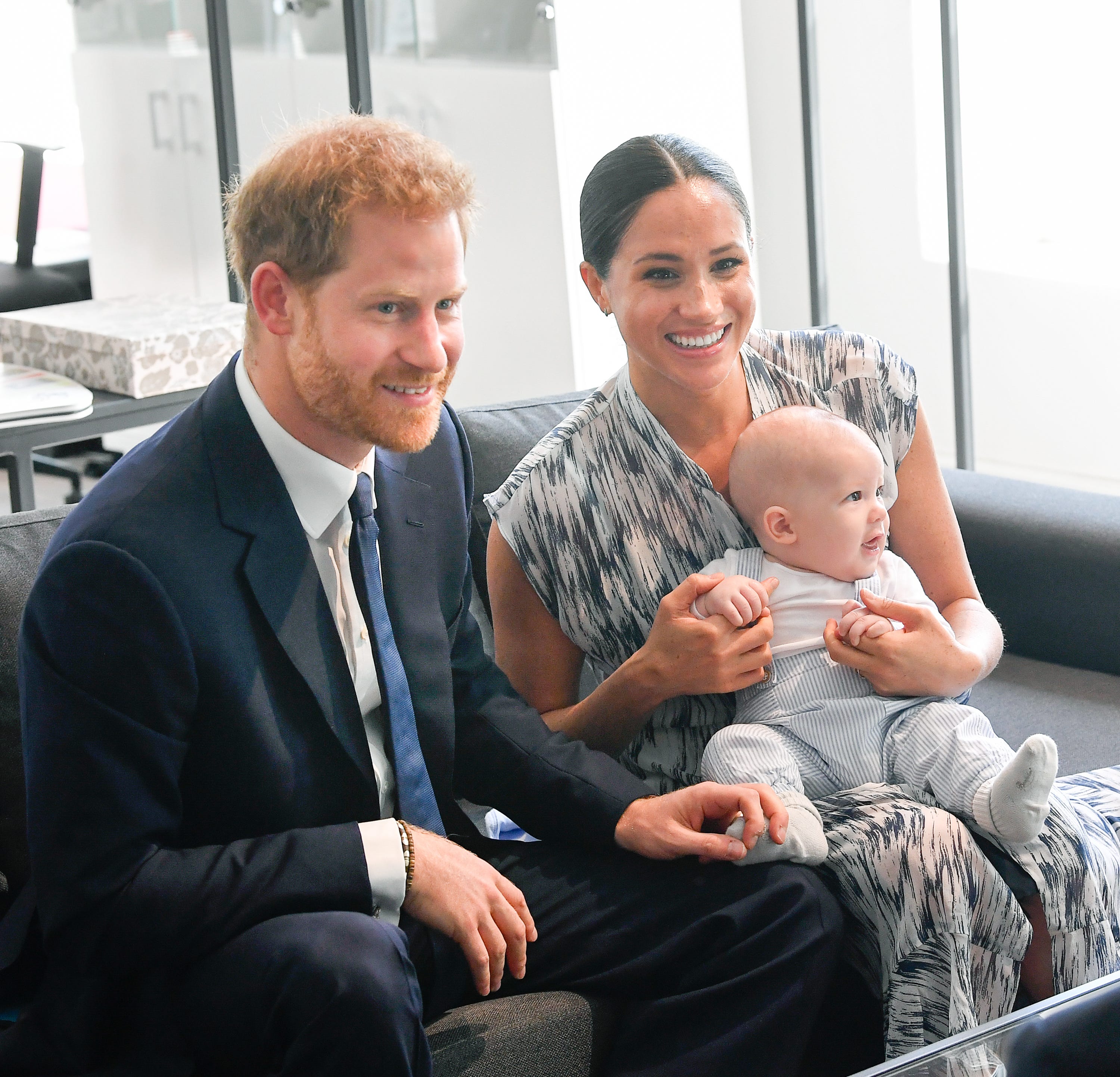 Prince Harry, dressed in a navy suit, sits on a couch next to Meghan Markle, who is wearing a gray and white patterned dress. Meghan is holding her son, Archie.