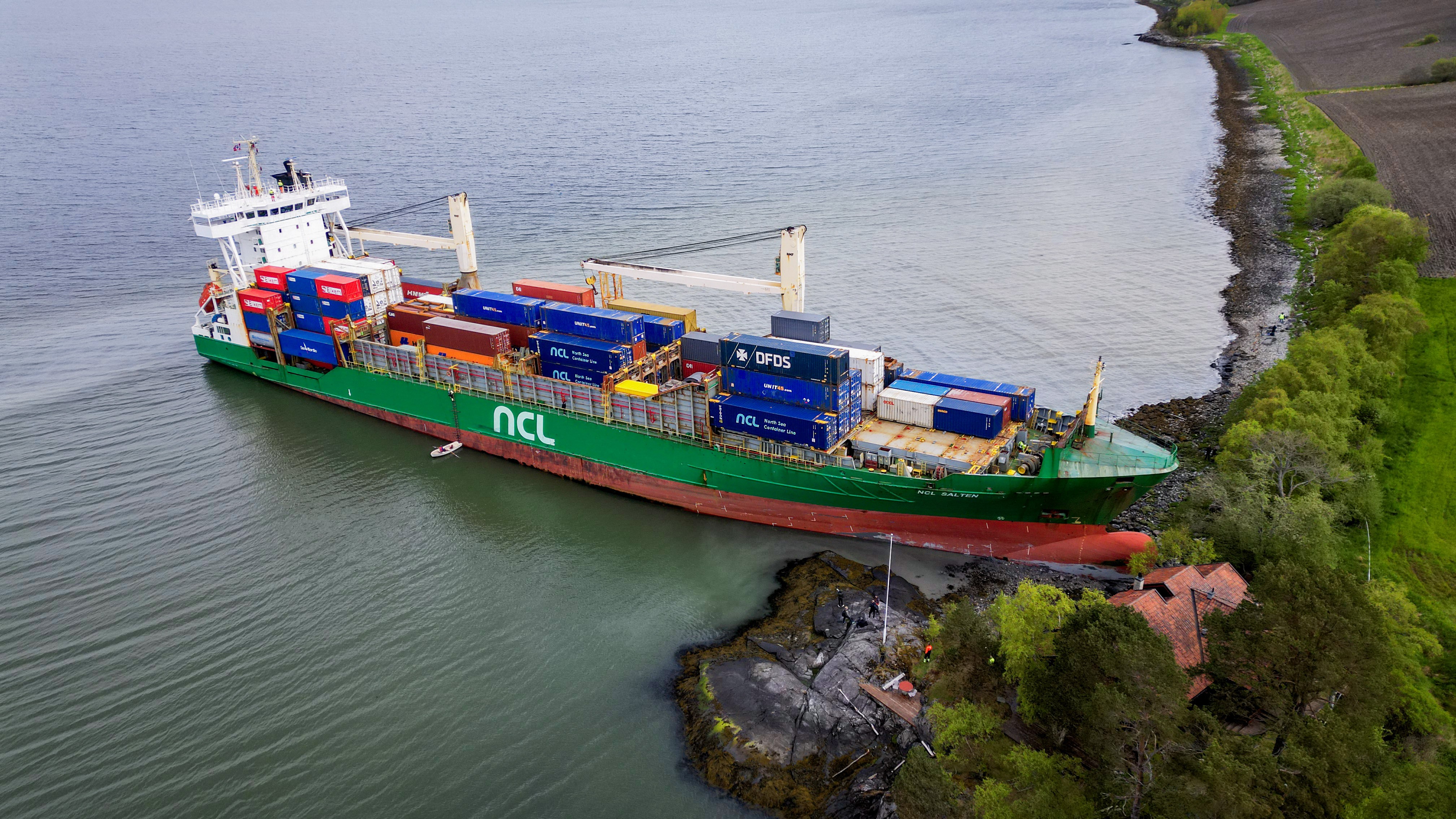 A container ship, its nose grounded on the shore, beside a house.