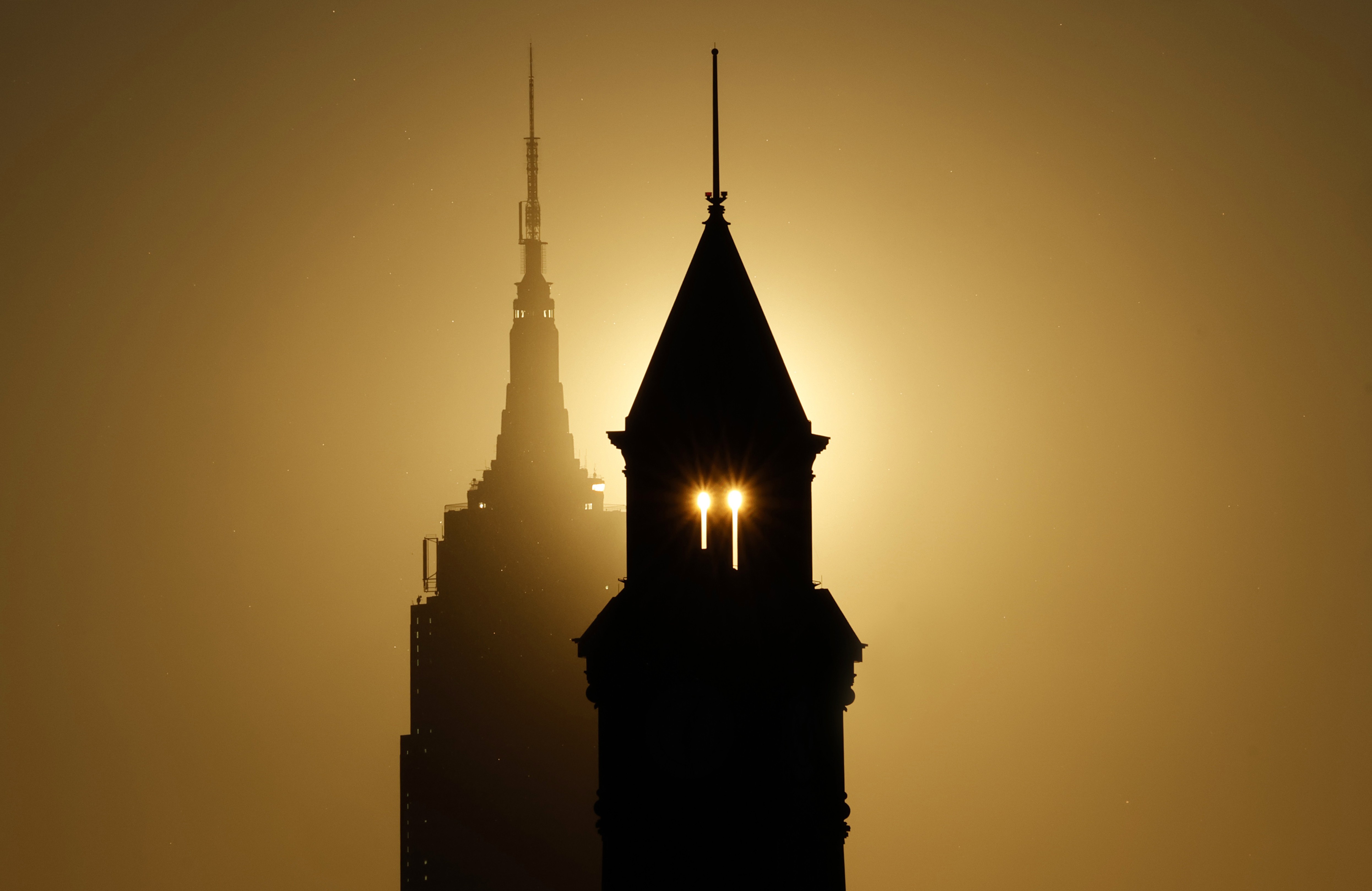 The sun rises behind the Empire State Building and another tower, silhouetting the buildings.