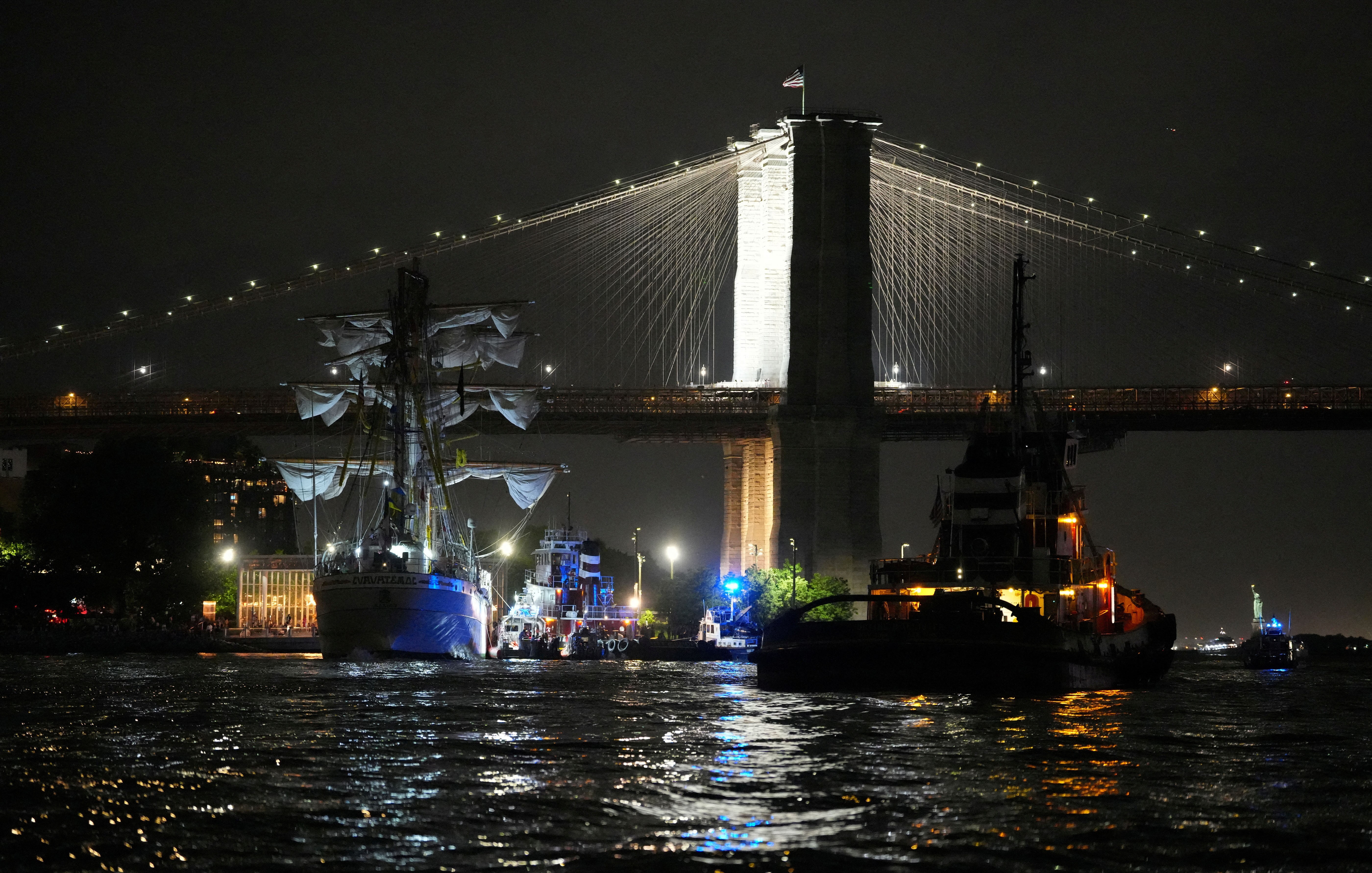 A tugboat and a damaged tall-masted ship float near the Brooklyn Bridge.