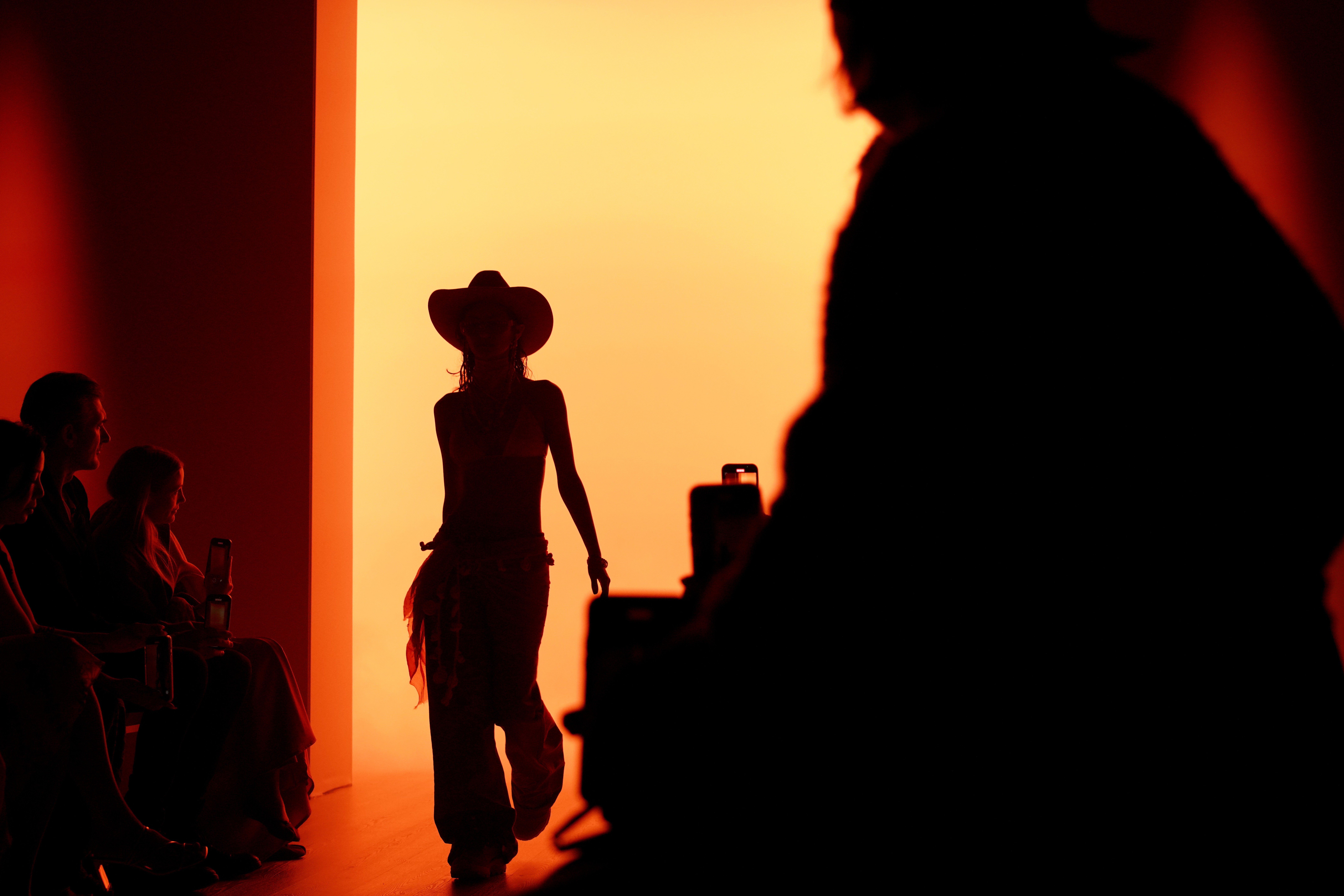 A model wearing a cowboy hat walks on a runway, seen in silhouette.