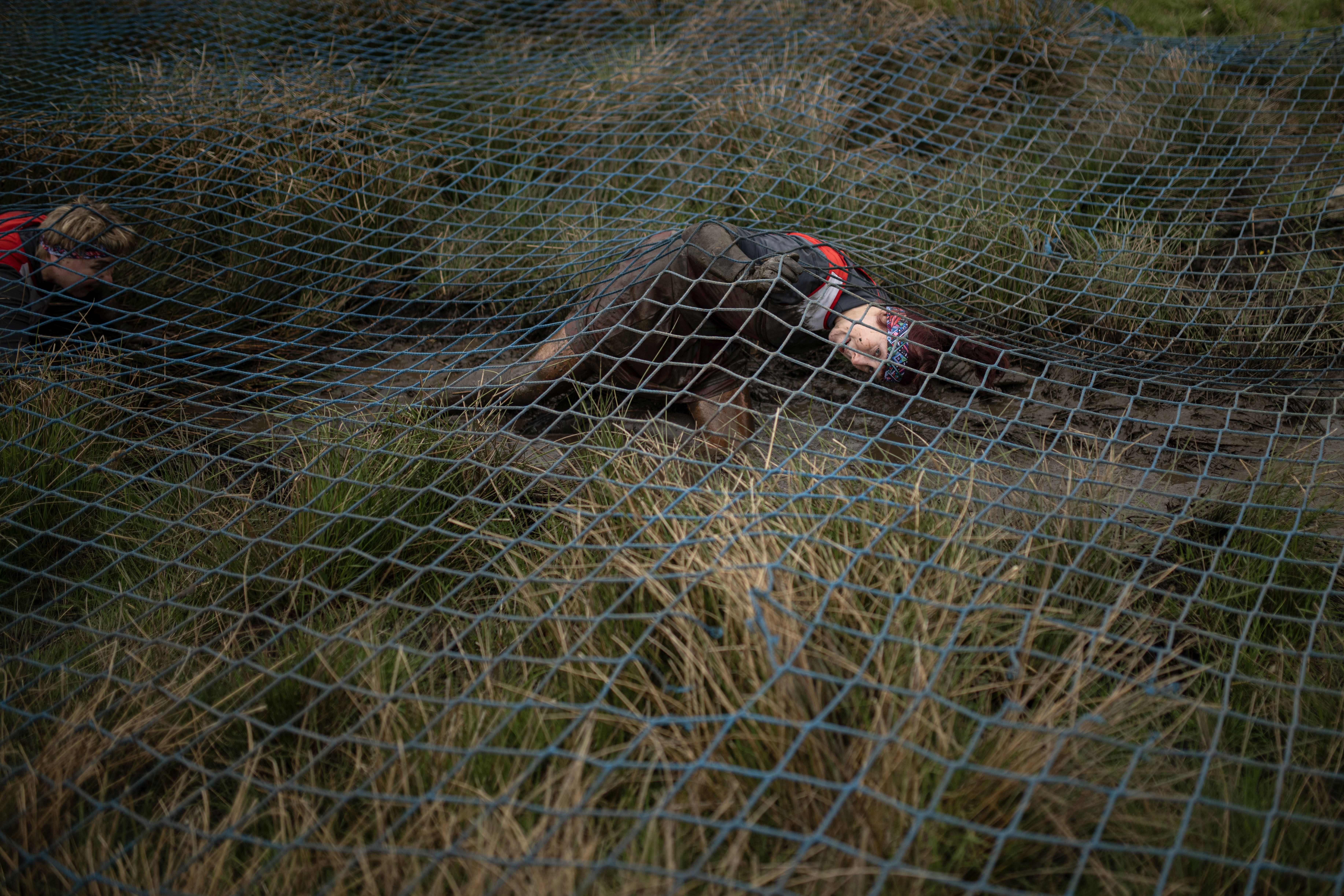 A competitor in a muddy obstacle race crawls through grass beneath netting.